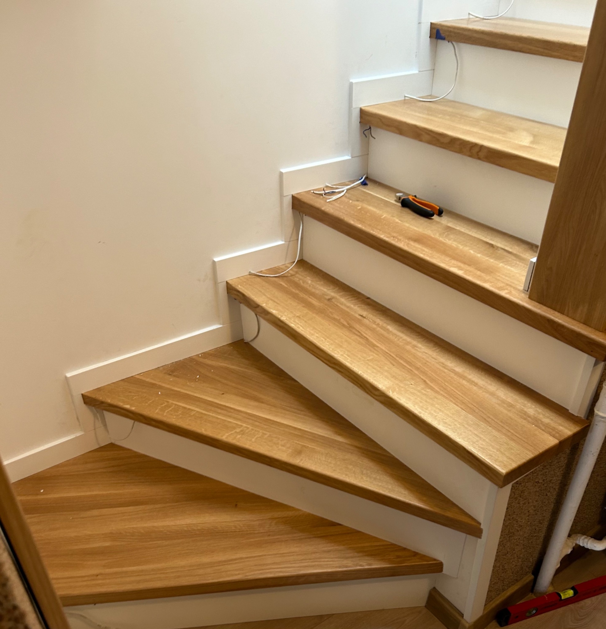 Montaż drewnianych schodów z białymi podstopnicami, widoczne przewody elektryczne i narzędzia na jednym ze stopni.