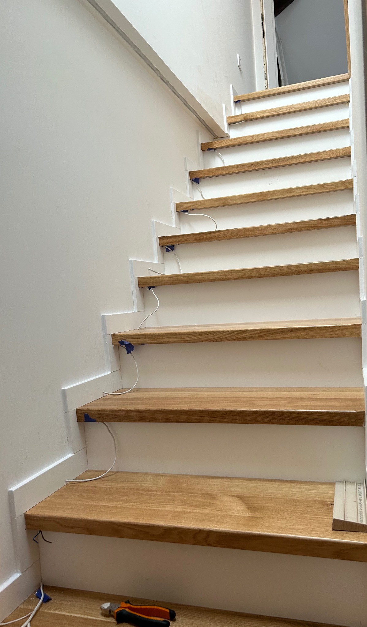 Montaż oświetlenia schodowego: surowe drewniane stopnie z białymi podstopnicami, widoczne przewody i niebieskie zabezpieczenia, narzędzia na podłodze.