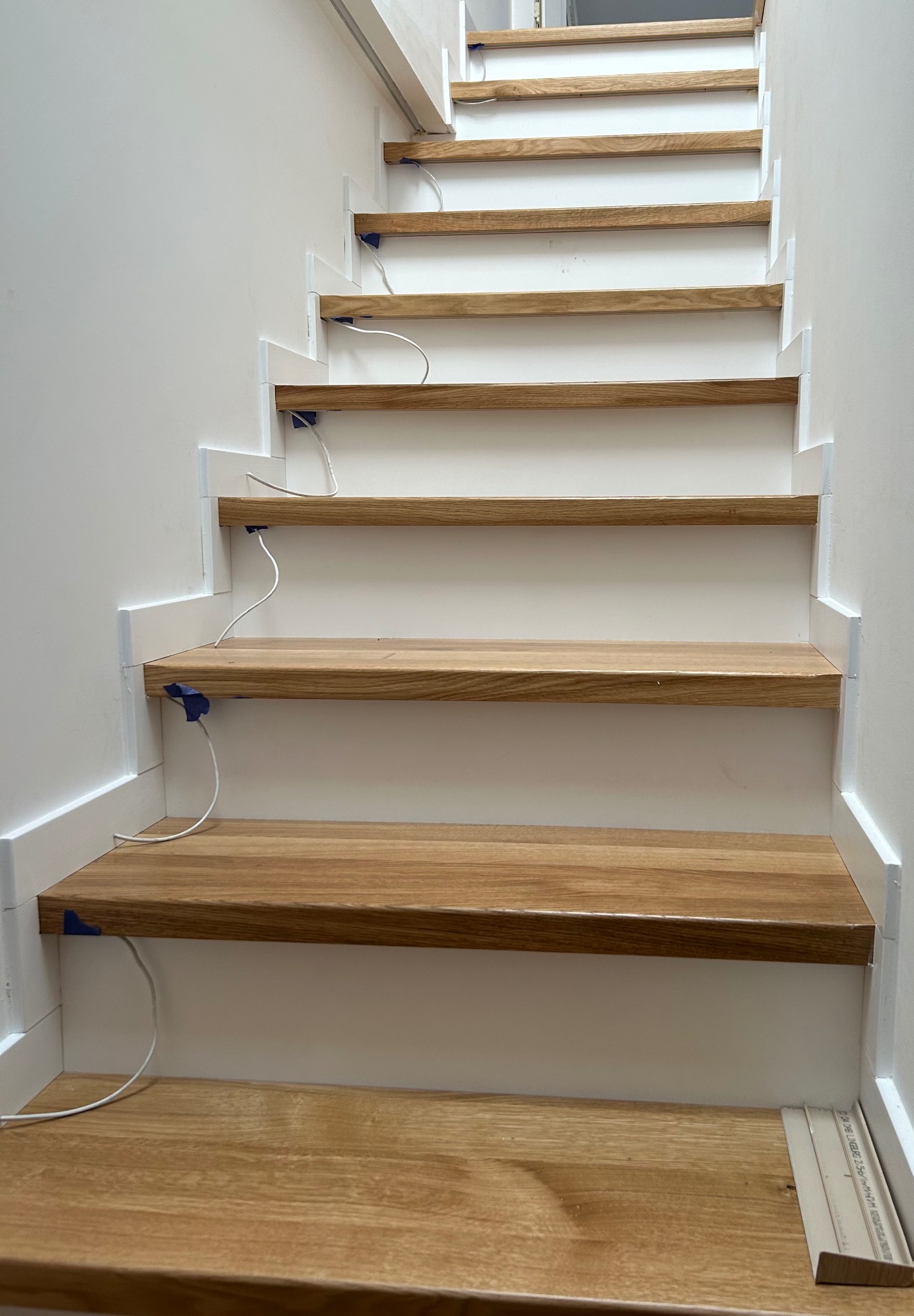 Fragment konstrukcji schodów z drewnianymi stopniami w trakcie montażu, zabezpieczone niebieską taśmą malarską i z widocznymi przewodami oświetleniowymi.