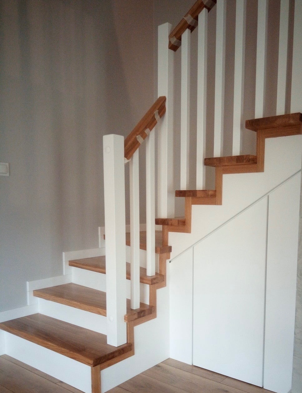 Nowoczesne schody z drewnianymi stopniami i białymi balustradami, zintegrowane z zabudową szafy wnękowej pod biegiem schodów.