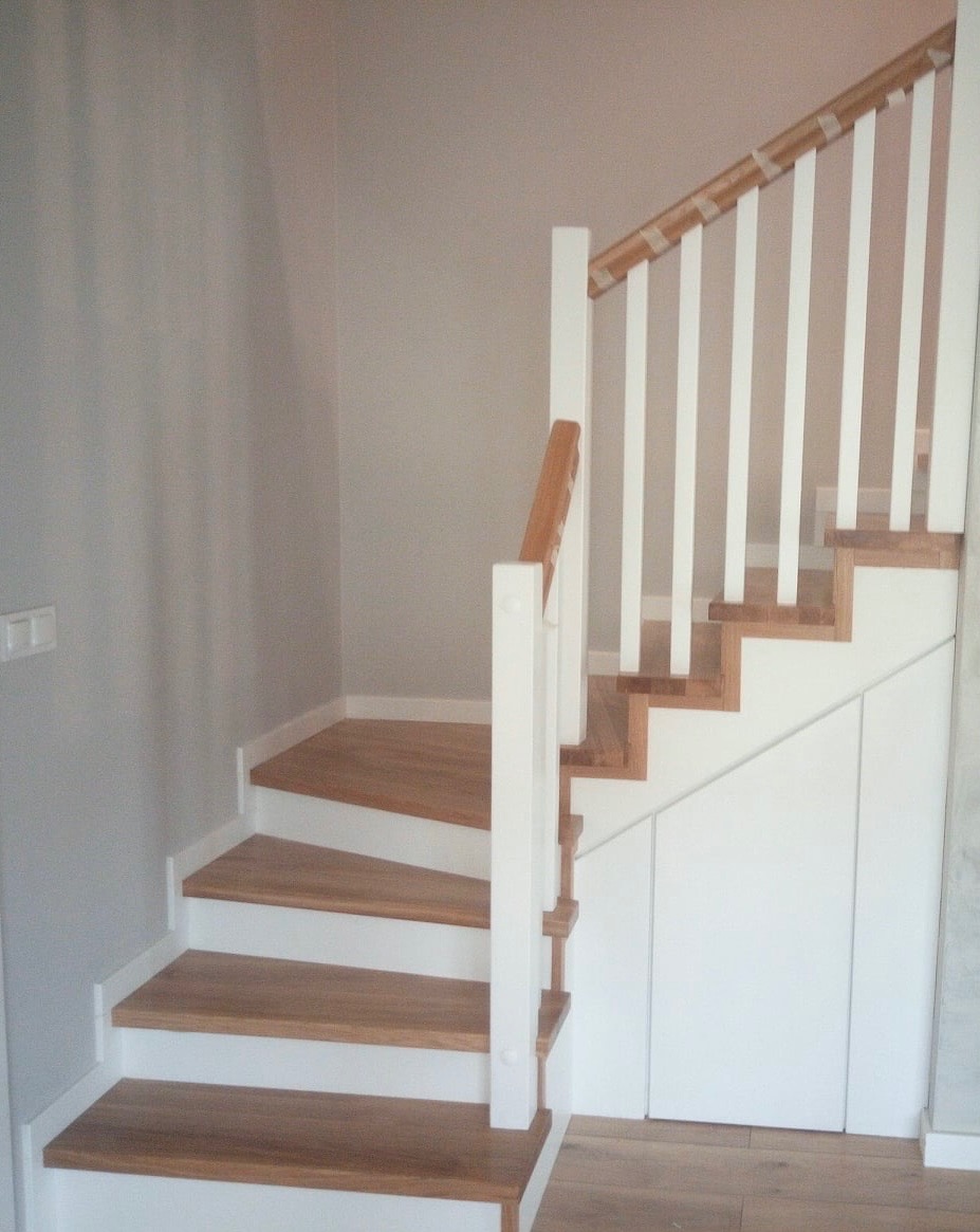 Jasne schody z drewnianymi stopniami i białą balustradą, z zabudową pod schodami w kolorze białym.