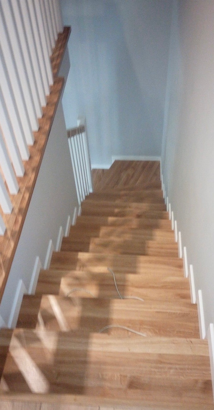 Widok z góry na drewniane schody z białymi balustradami, częściowo zacienione, z widocznymi kablami na stopniach, prowadzące w dół.