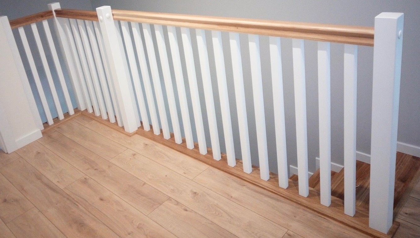 Nowoczesna balustrada schodowa z białymi pionowymi szczeblami i drewnianym poręczą, zamontowana na drewnianej podłodze.