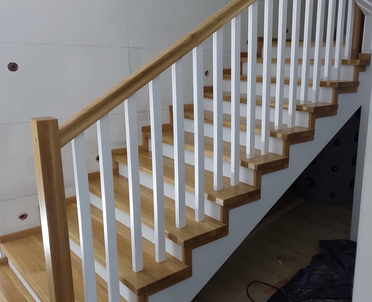 Drewniane schody z białymi balustradami, widoczne na tle surowej ściany w trakcie wykończenia, z elementami instalacji elektrycznej.