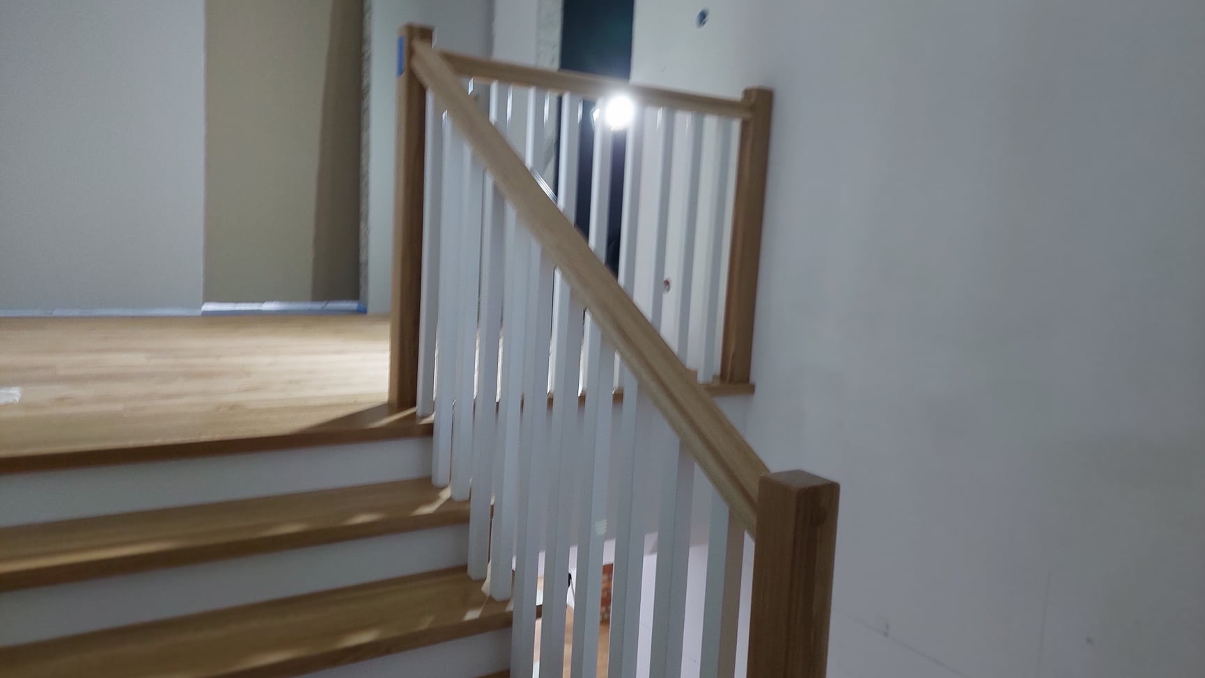 Fragment drewnianych schodów z białą balustradą w trakcie montażu, widoczne niedomalowane ściany i zabezpieczenia podłogi.