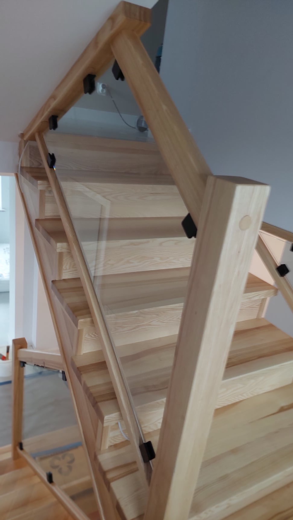 Jasne, drewniane schody z nowoczesną szklaną balustradą, mocowaną czarnymi uchwytami, widok z dołu w kierunku góry schodów.