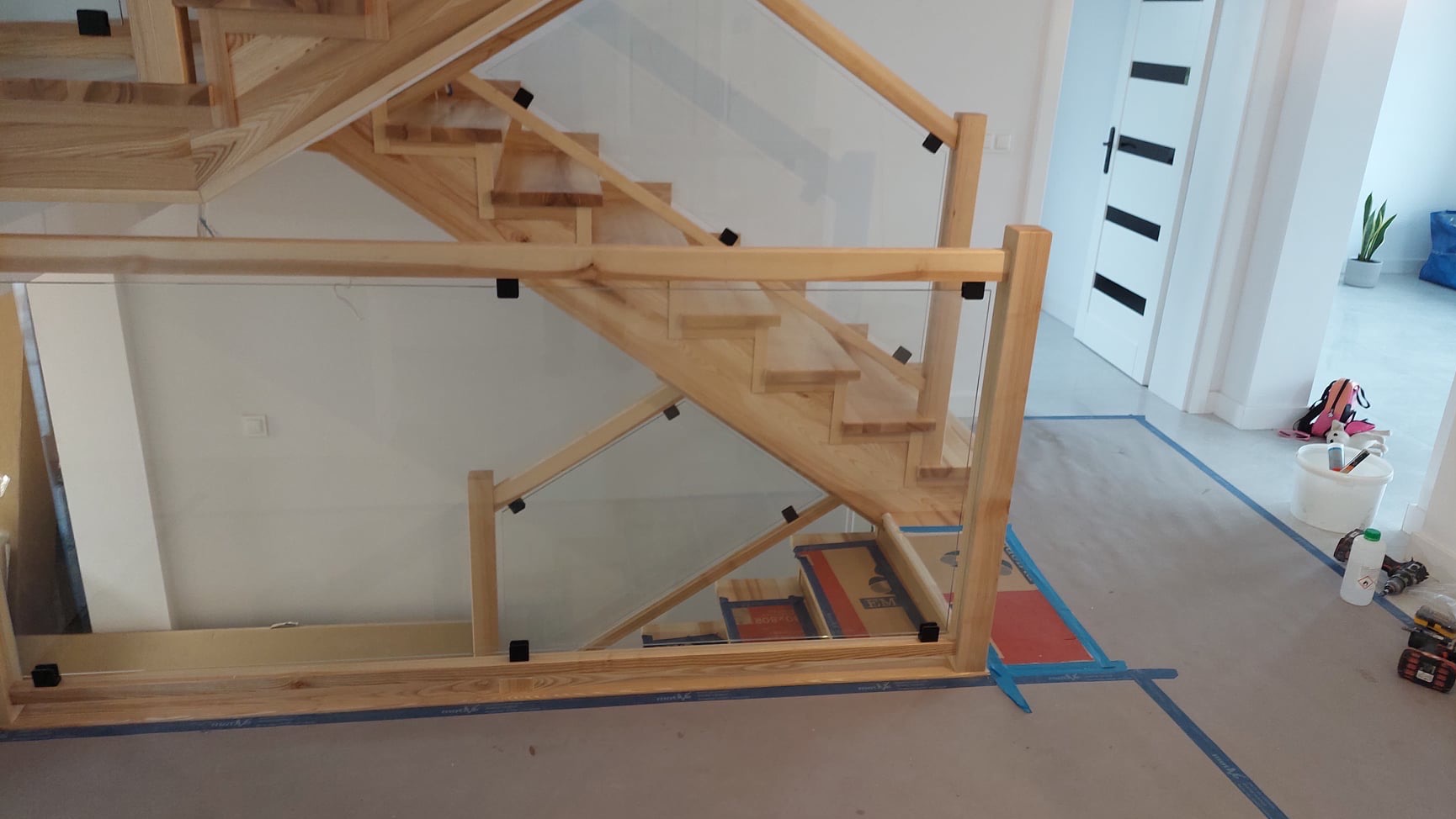 Jasne drewniane schody z balustradą ze szkła hartowanego, widoczne elementy montażowe w kolorze czarnym, podłoga zabezpieczona folią malarską, w tle białe drzwi i roślina doniczkowa.