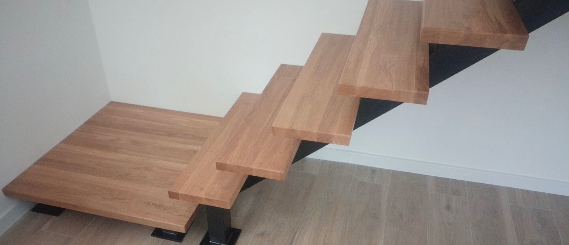 Nowoczesne schody z drewnianymi stopniami i czarną metalową konstrukcją wsporczą przy białej ścianie i podłodze z paneli.