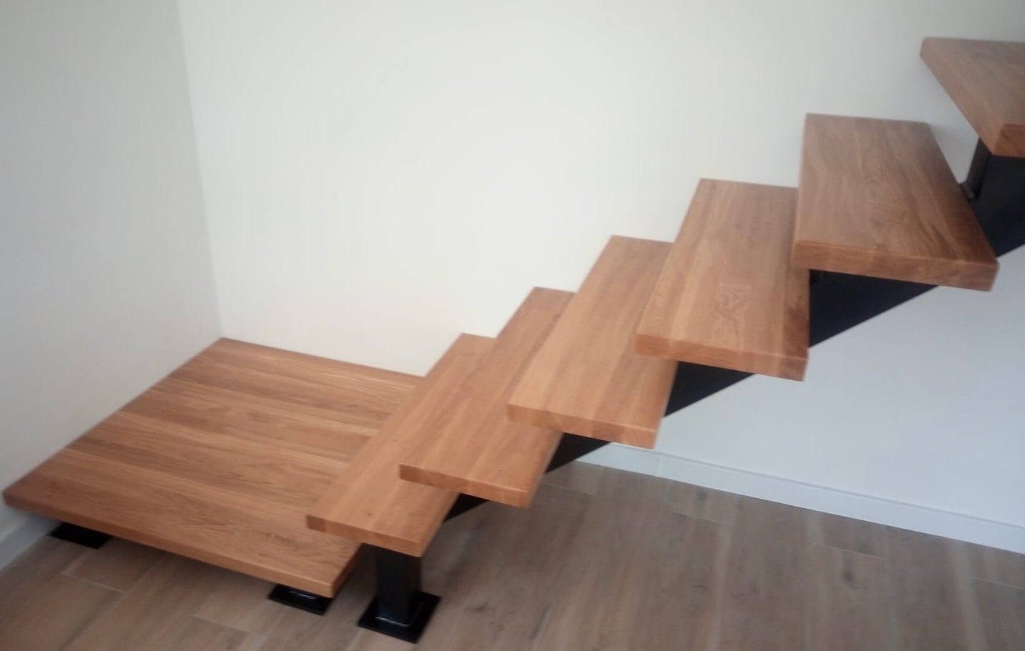 Nowoczesne schody z drewna dębowego, wspornikowe, z czarnymi metalowymi elementami mocującymi, widoczne z dołu, na tle białej ściany i drewnopodobnej podłogi.