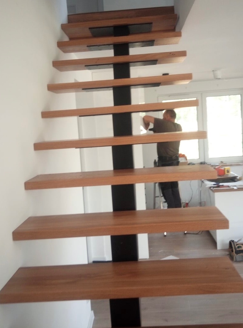 Montaż schodów drewnianych z centralną, czarną belką nośną; widoczny pracownik podczas instalacji w jasnym wnętrzu.