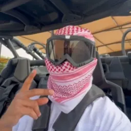 Mężczyzna w kefii i goglach ochronnych, siedzący w pojeździe typu buggy, z zapiętymi pasami bezpieczeństwa, wykonuje gest palcami.