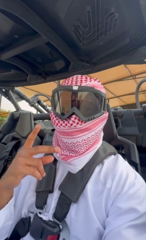 Mężczyzna w kefii i goglach ochronnych, siedzący w pojeździe typu buggy, z zapiętymi pasami bezpieczeństwa, wykonuje gest palcami.