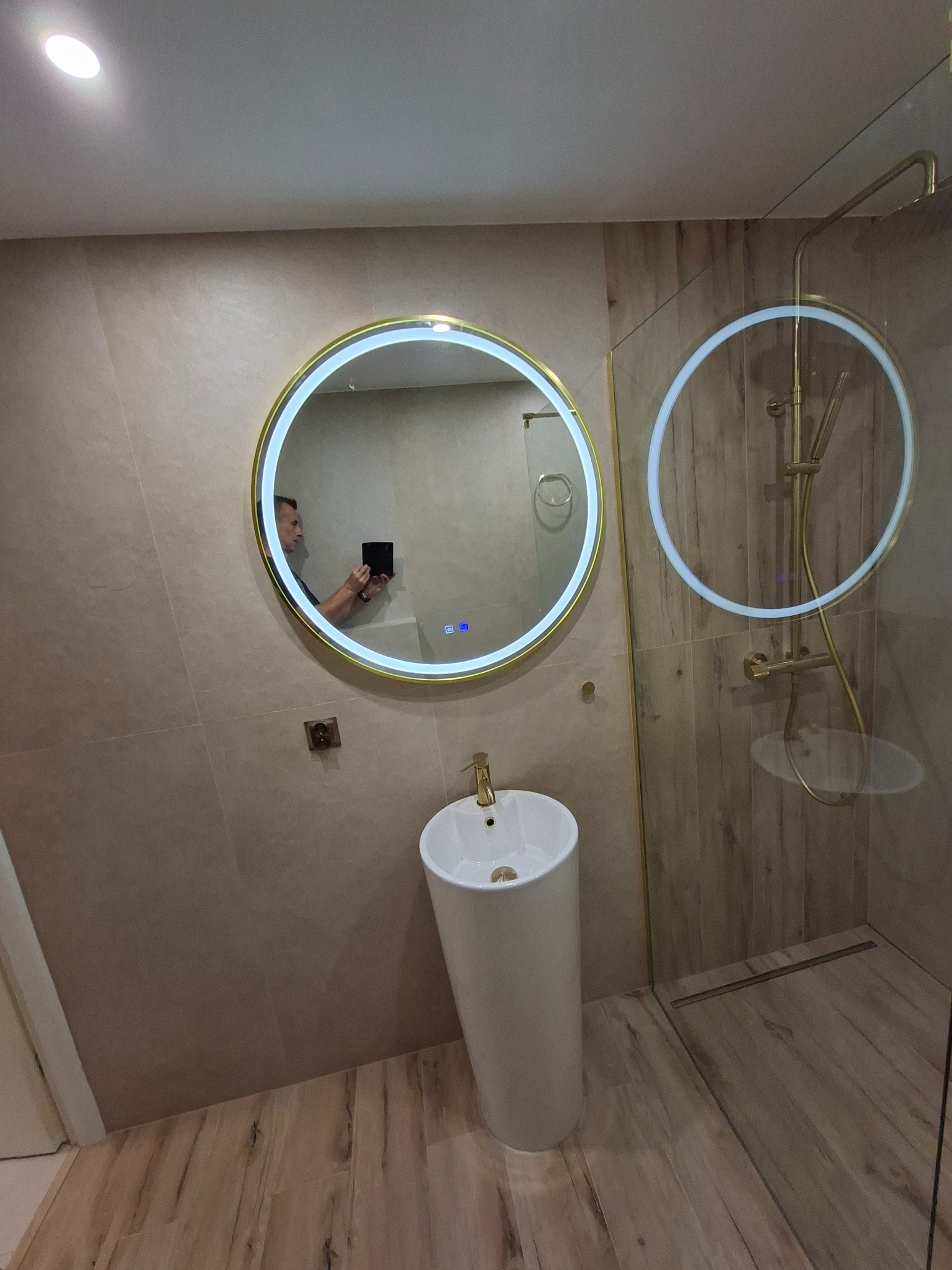 Wykończona łazienka z okrągłym lustrem LED, białą umywalką na postumencie i kabiną prysznicową ze złotą armaturą. Beżowe płytki imitujące drewno na ścianach i podłodze.
