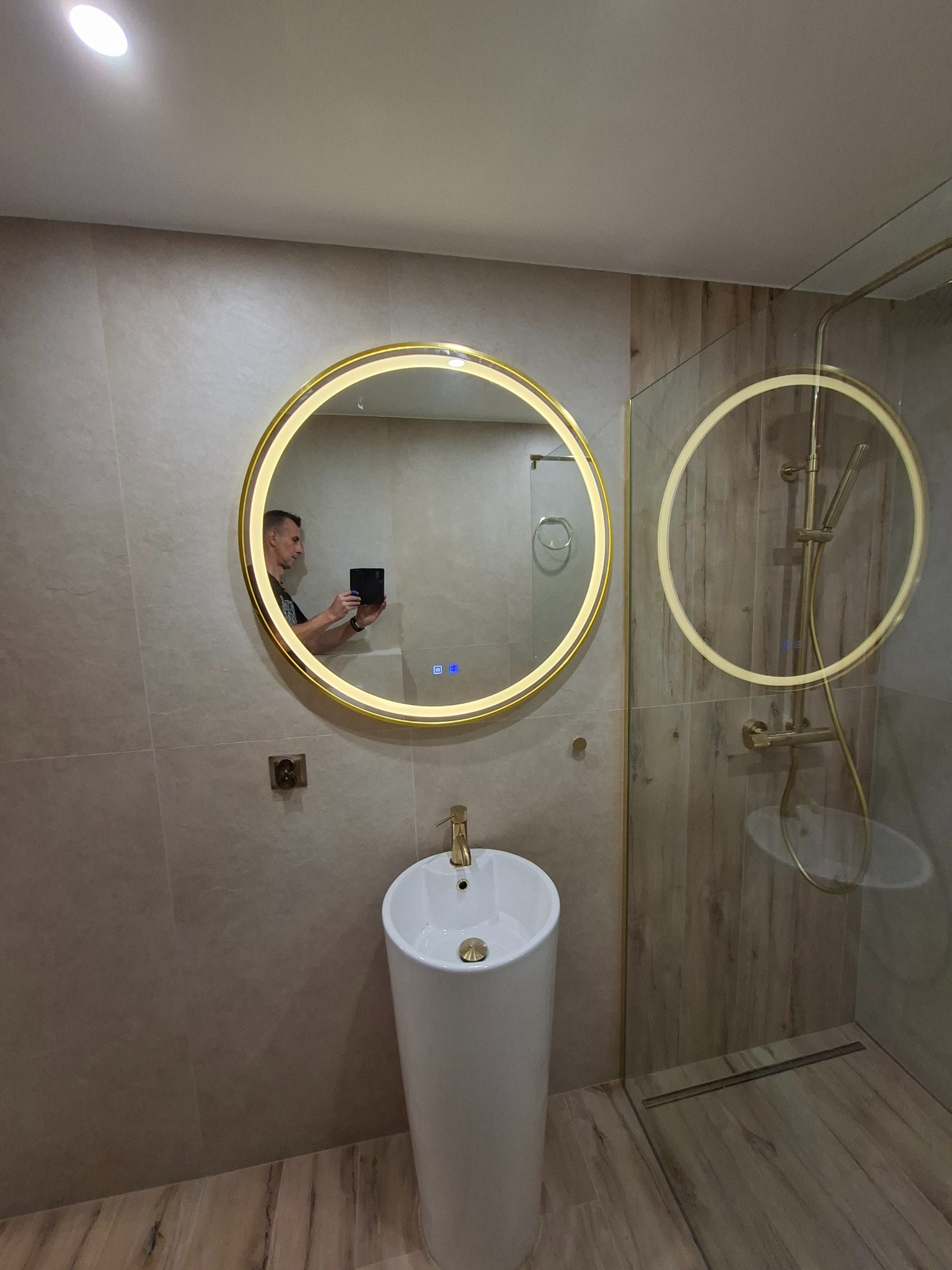 Nowoczesna łazienka z okrągłym lustrem LED, białą umywalką kolumnową i kabiną prysznicową ze złotymi akcentami. Widoczne płytki imitujące drewno.