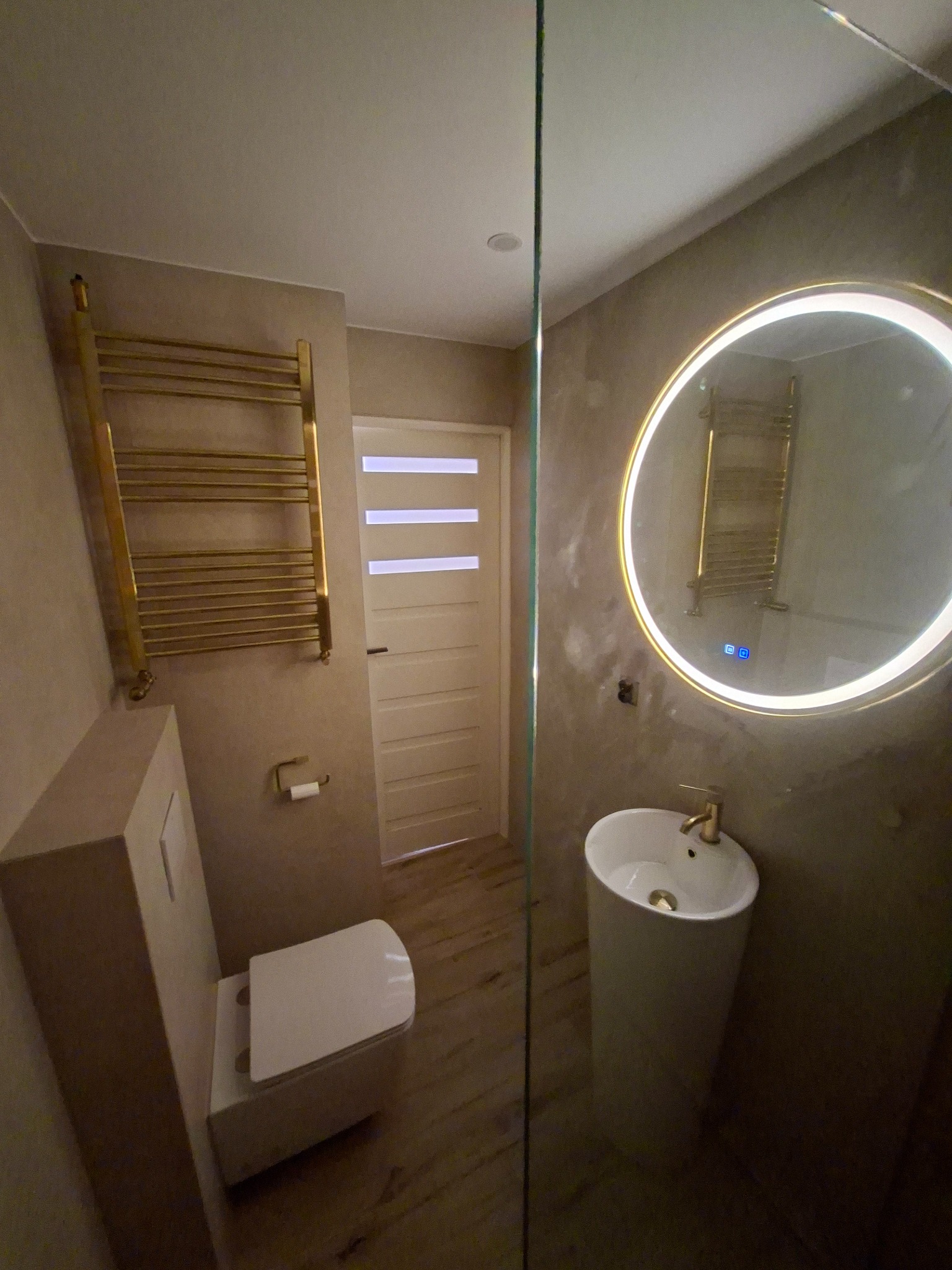 Nowoczesna łazienka z okrągłym lustrem LED, złotym grzejnikiem drabinkowym i minimalistyczną umywalką na postumencie. Beżowe ściany i drewnopodobna podłoga.