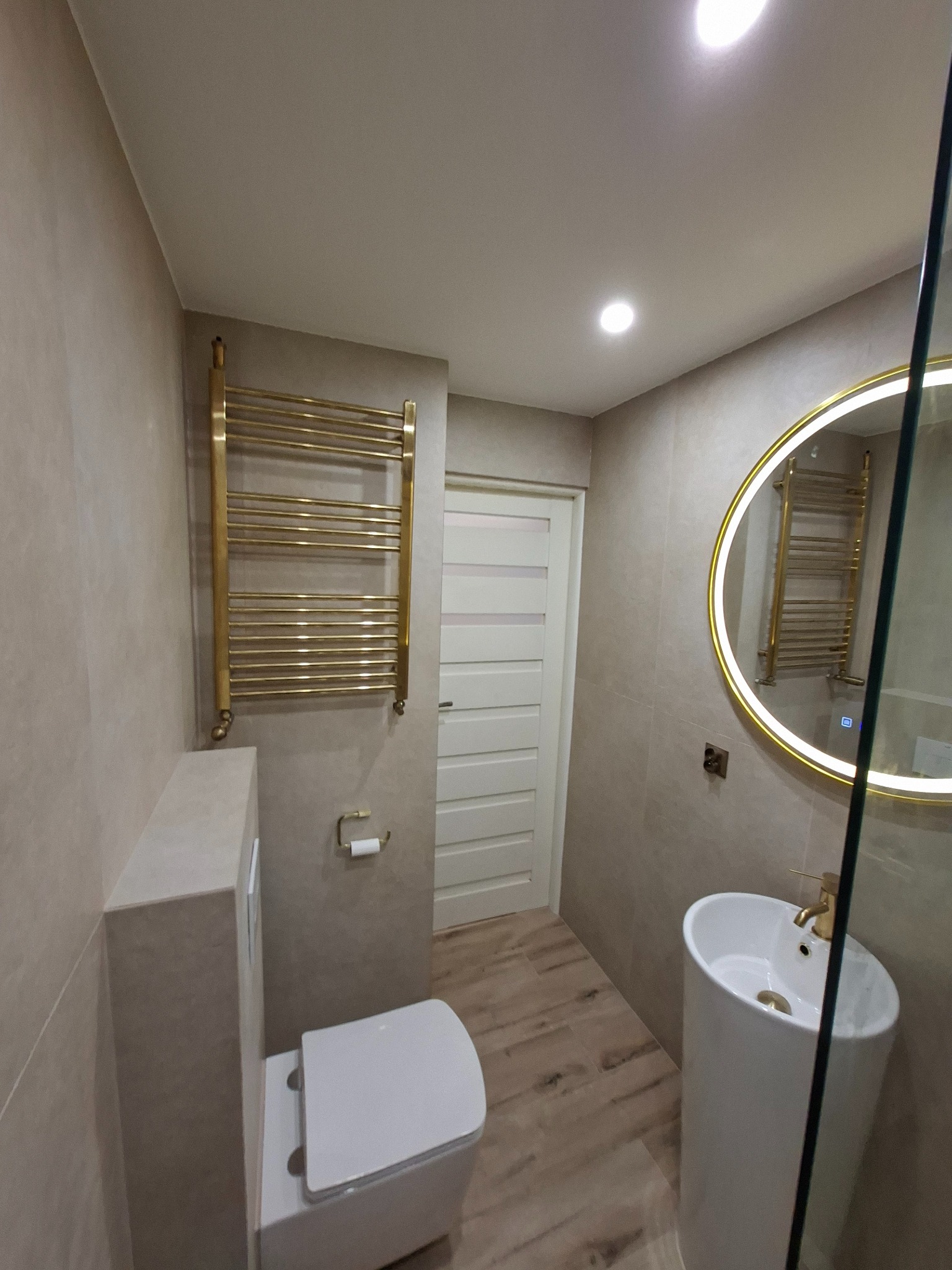 Nowoczesna łazienka w beżowych tonacjach z złotymi akcentami: grzejnik drabinkowy, okrągłe lustro z podświetleniem LED i wolnostojąca umywalka.