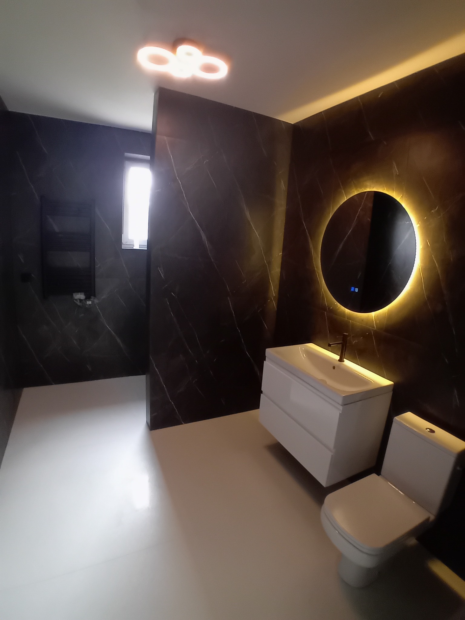 Nowoczesna łazienka z czarnymi marmurowymi ścianami, białymi sanitariatami i okrągłym lustrem z podświetleniem LED. Minimalistyczny design, ciepłe oświetlenie.