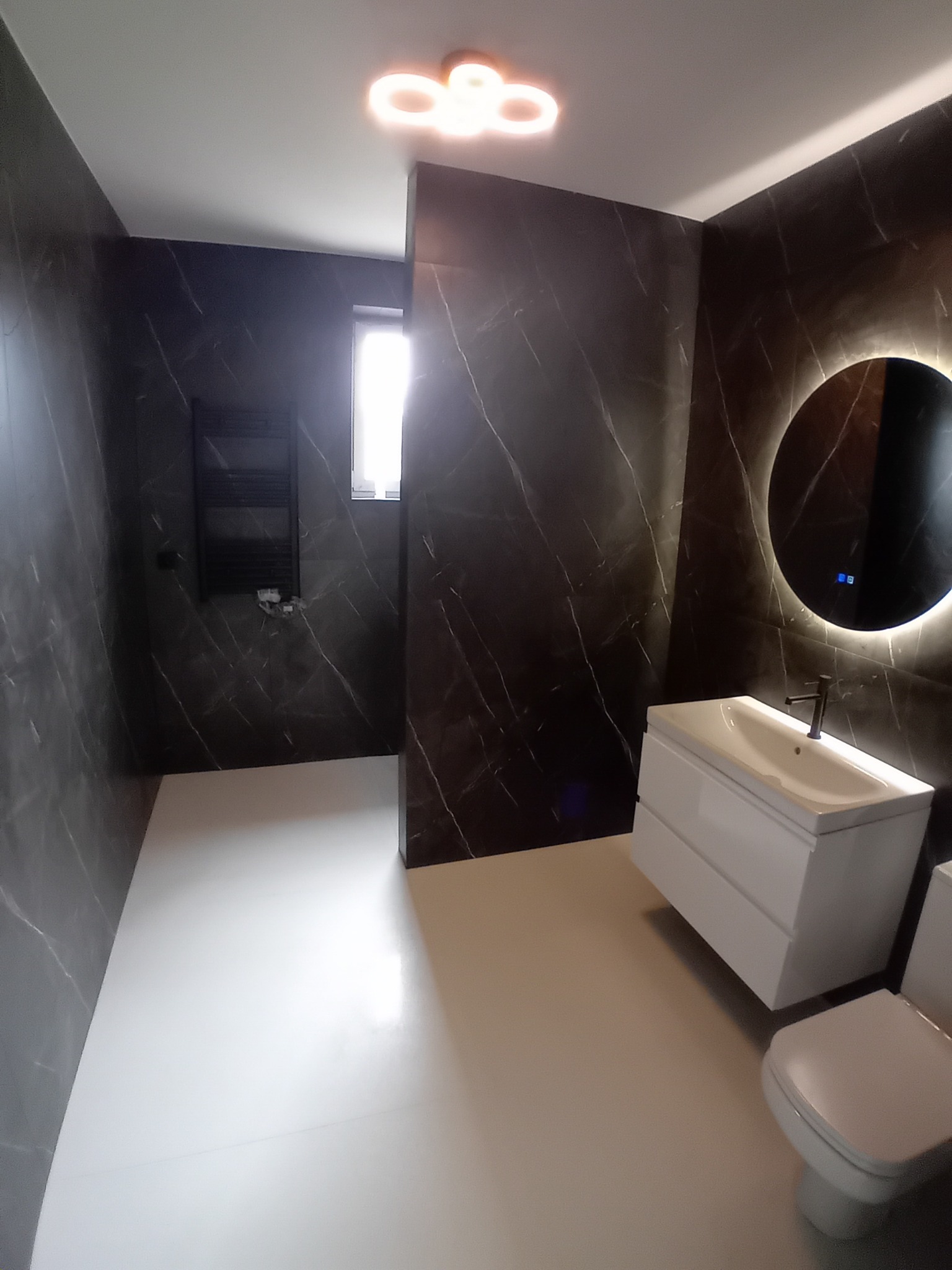 Nowoczesna łazienka z czarnymi płytkami imitującymi marmur, białymi sanitariatami i okrągłym lustrem z podświetleniem LED. Minimalistyczny design, czyste linie.