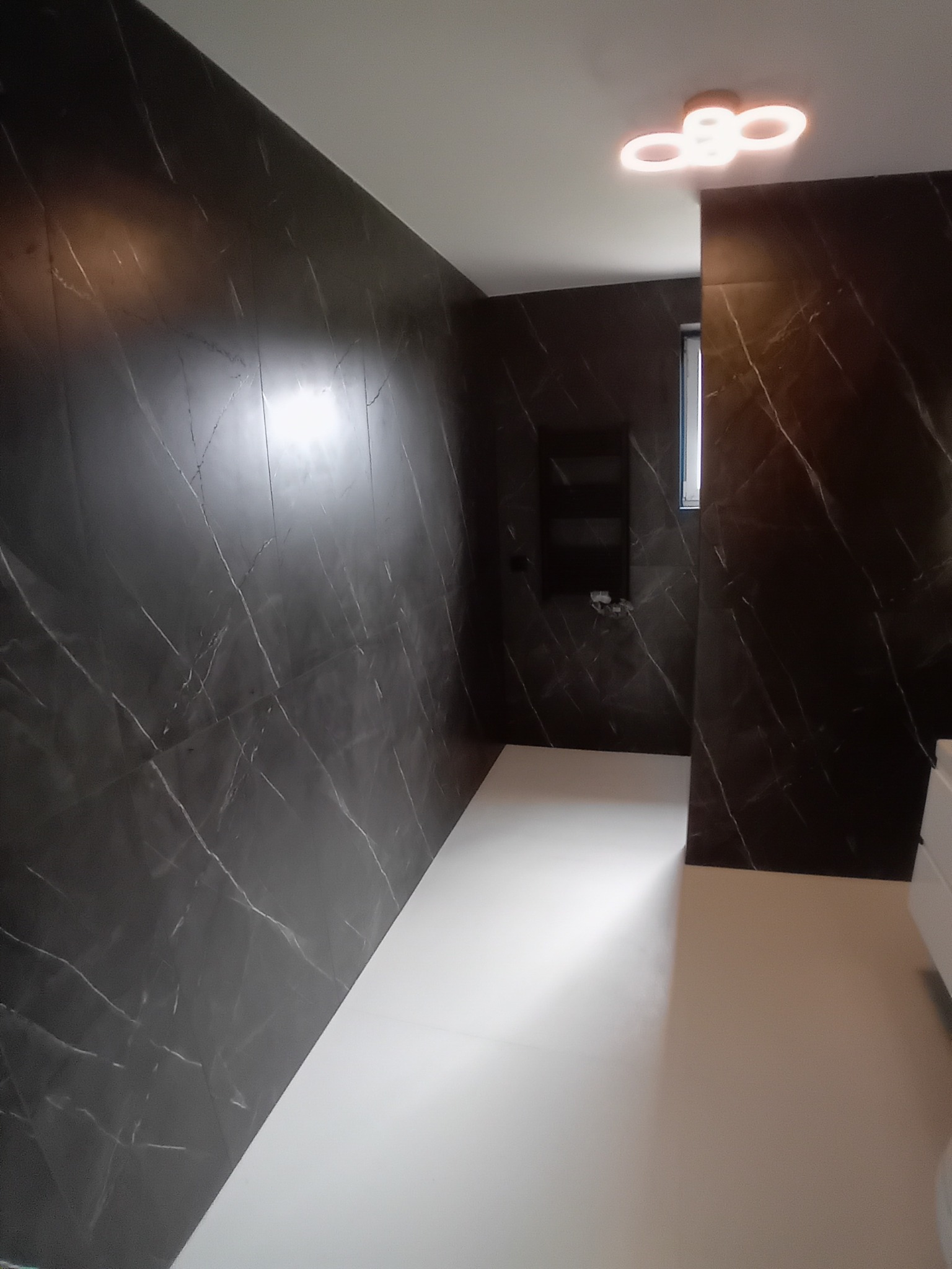 Nowoczesna łazienka z czarnymi płytkami imitującymi marmur i białymi podłogami. Widoczna zabudowa wnęki i designerska lampa sufitowa.