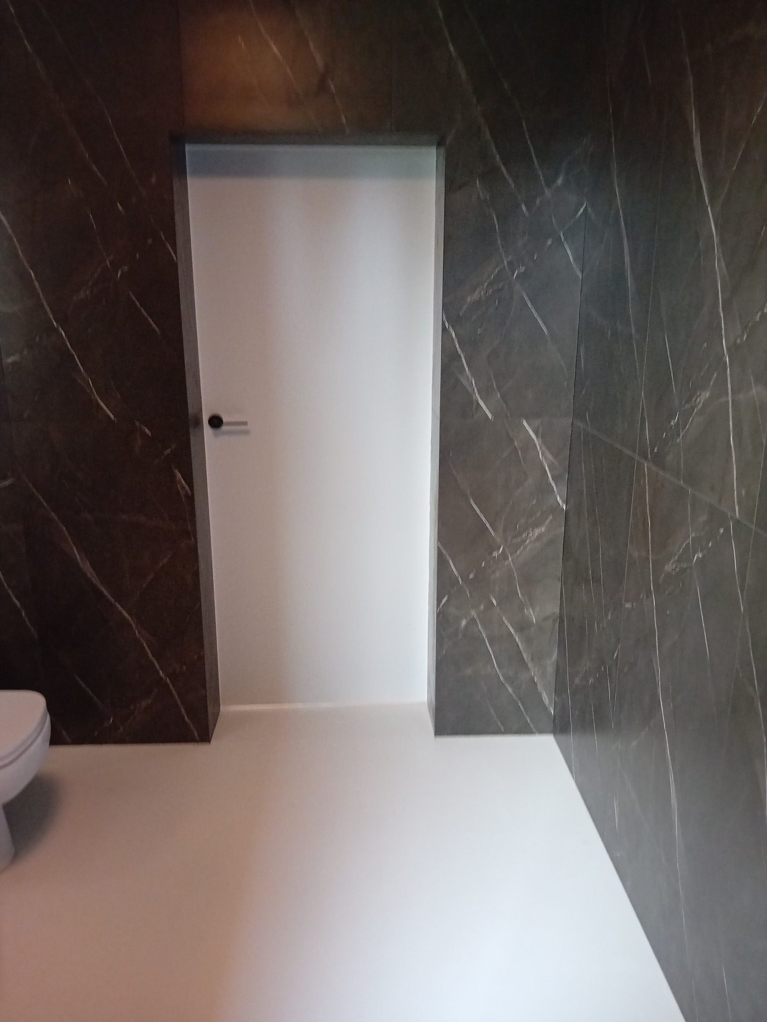 Nowoczesna łazienka z marmurowymi płytkami w odcieniach szarości i brązu, białą podłogą i drzwiami. Minimalistyczny design, widoczny fragment toalety.