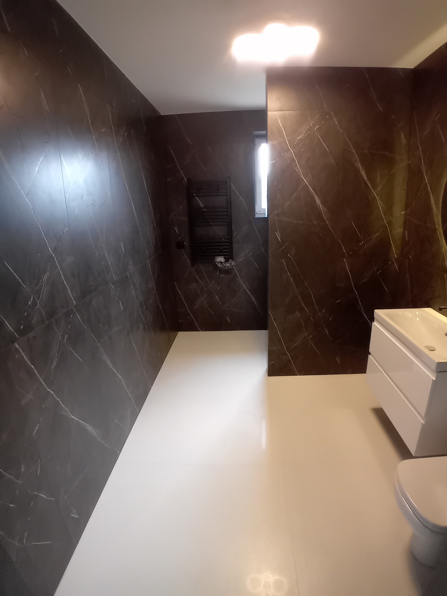 Nowoczesna łazienka z ciemnymi płytkami imitującymi marmur na ścianach i jasną podłogą. Widoczna umywalka z szafką i grzejnik drabinkowy.