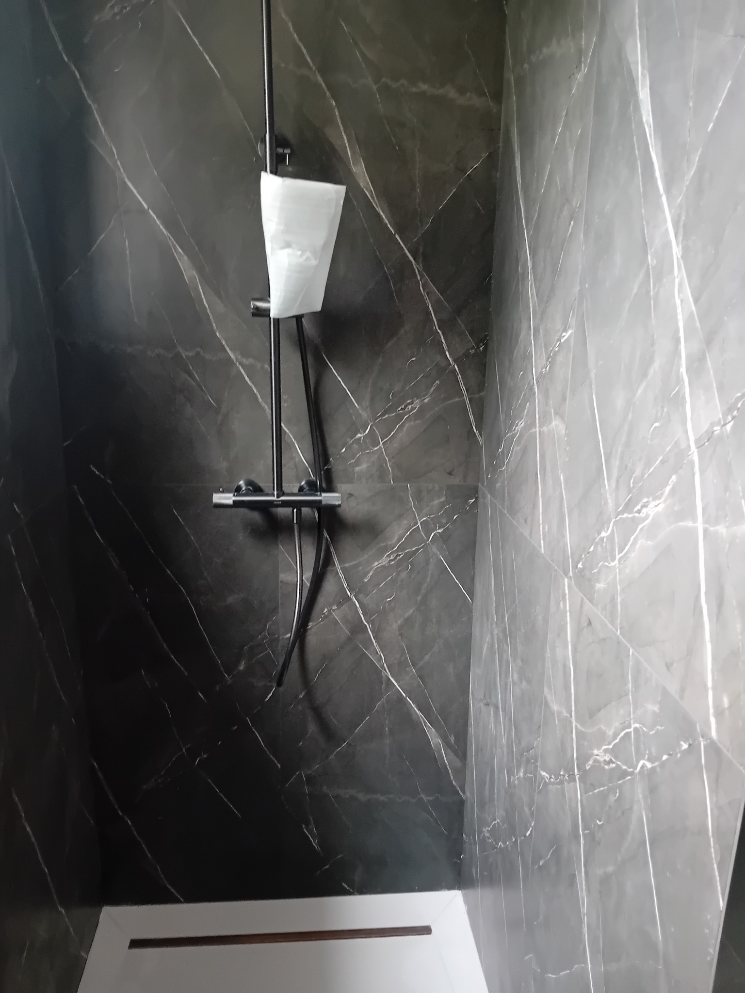 Nowoczesna kabina prysznicowa z czarnymi płytkami imitującymi marmur i białą brodzikiem. Bateria prysznicowa w folii ochronnej.