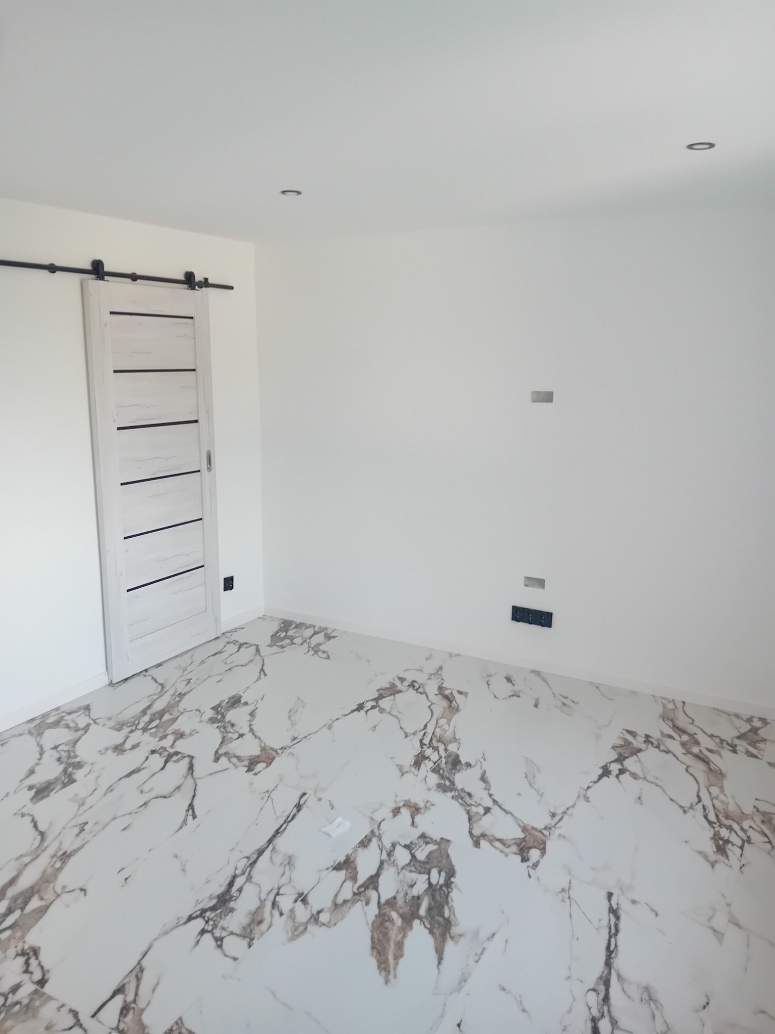 Jasne wnętrze po remoncie: marmurowe płytki podłogowe z brązowymi żyłami, białe ściany z gniazdkami i przesuwnymi drzwiami w stylu stodoły. Minimalistyczny design.