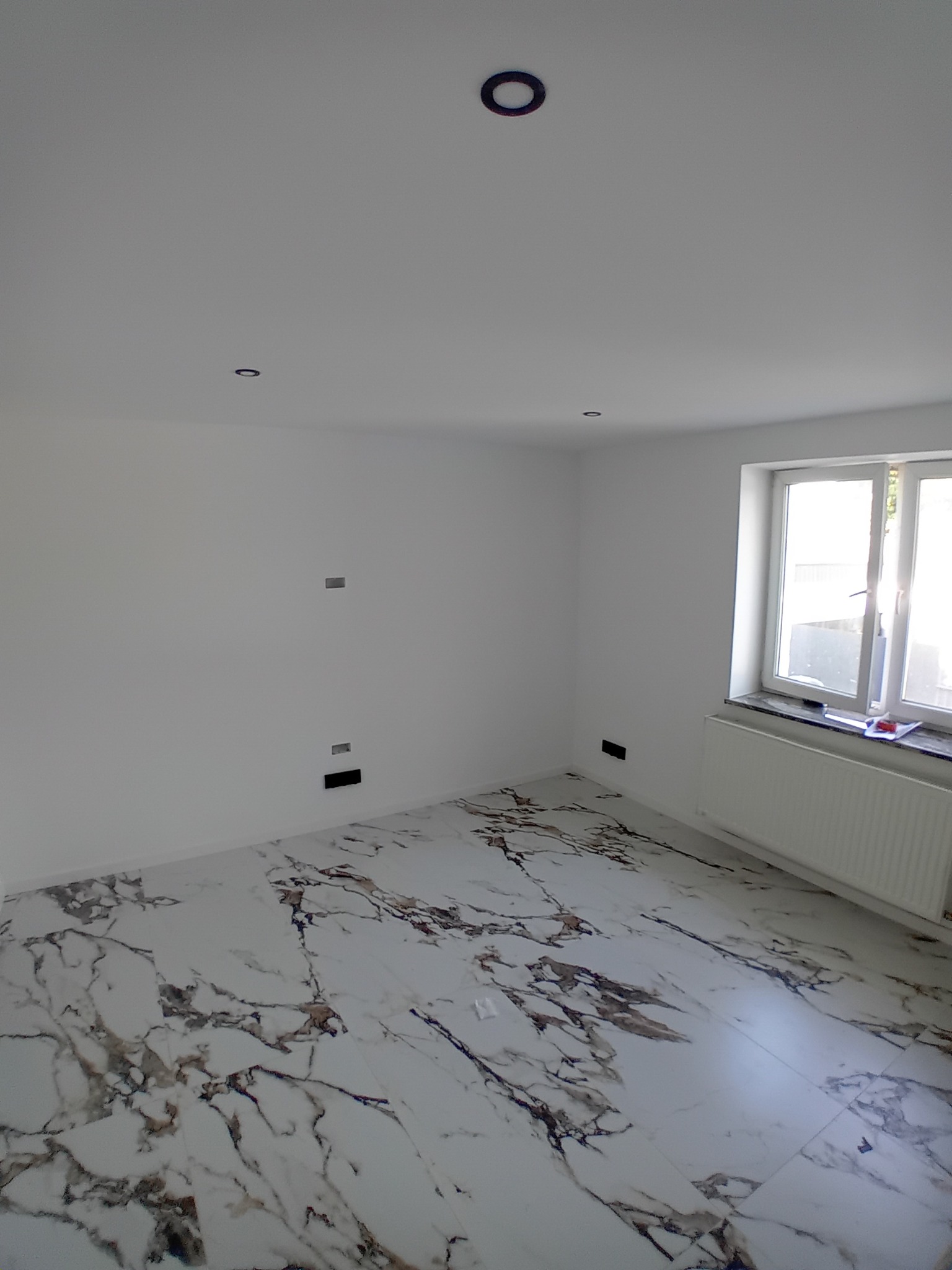 Białe wnętrze po remoncie: marmurowe płytki podłogowe, świeżo pomalowane ściany z gniazdkami, okno z grzejnikiem. Minimalistyczny design, widok z perspektywy podłogi.