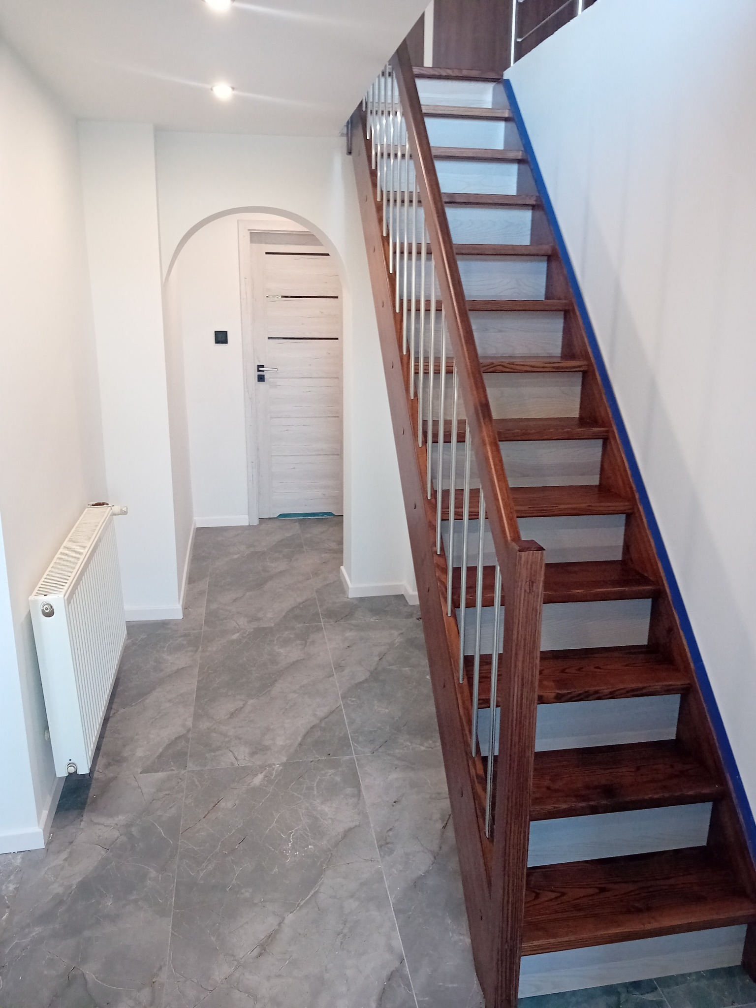 Nowoczesne schody drewniane z metalowymi barierkami w jasnym wnętrzu z szarą podłogą imitującą marmur. Widoczny grzejnik i drzwi w łuku.