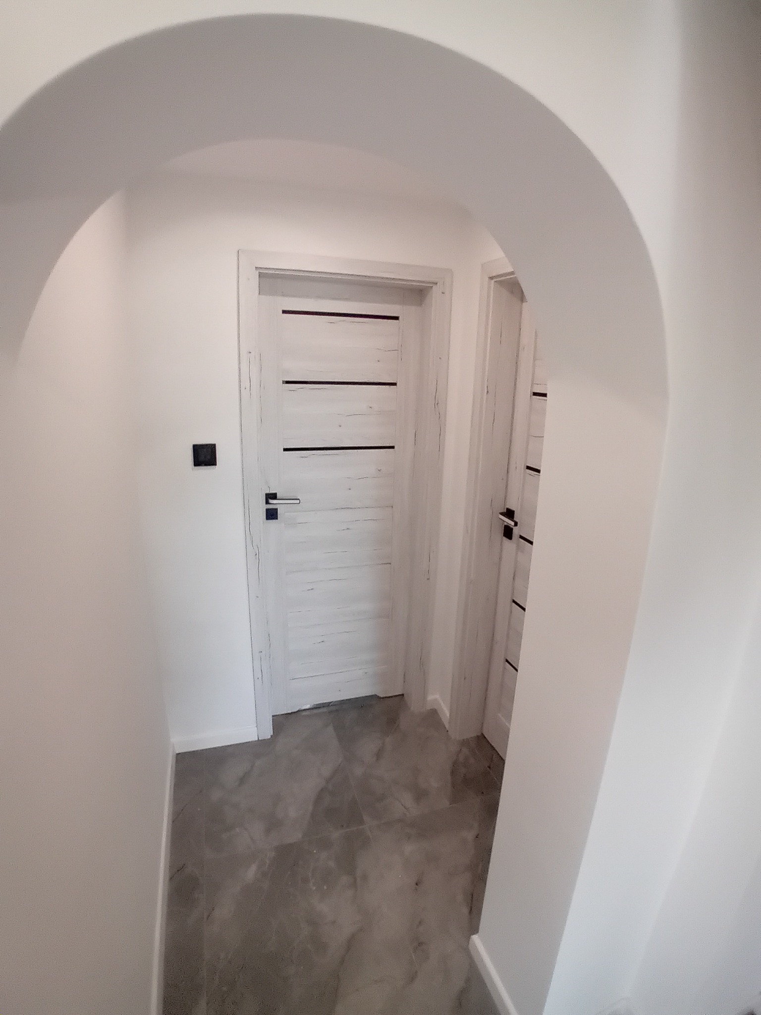 Białe wnętrze z łukowym przejściem, szarą marmurową podłogą i dwiema drewnianymi, bielonymi drzwiami z poziomymi zdobieniami. Minimalistyczny design.