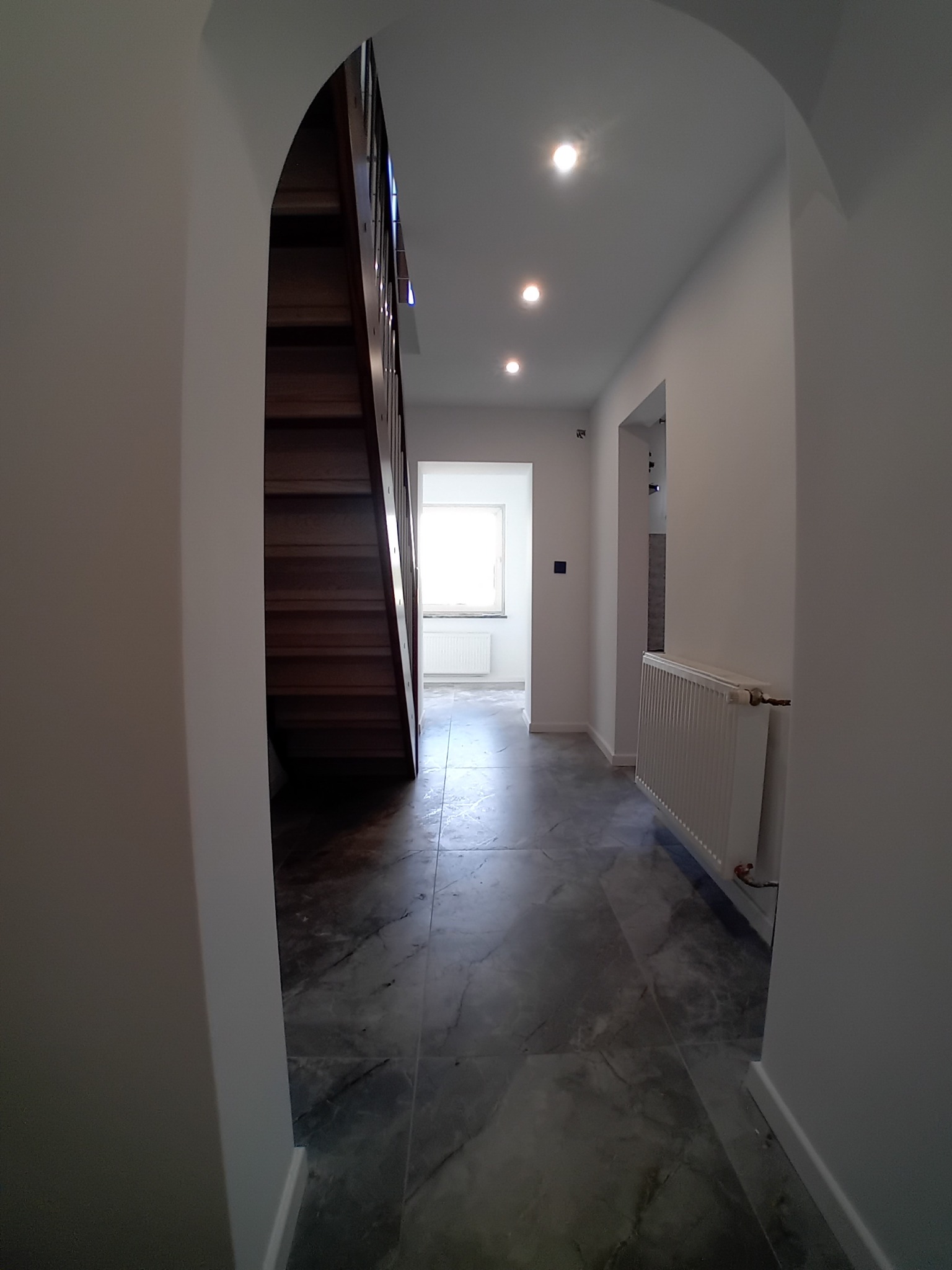 Wykończony korytarz z marmurową posadzką i drewnianymi schodami. Białe ściany, łukowe przejście, grzejnik. Widok na okno w oddali. Nowoczesne oświetlenie sufitowe.