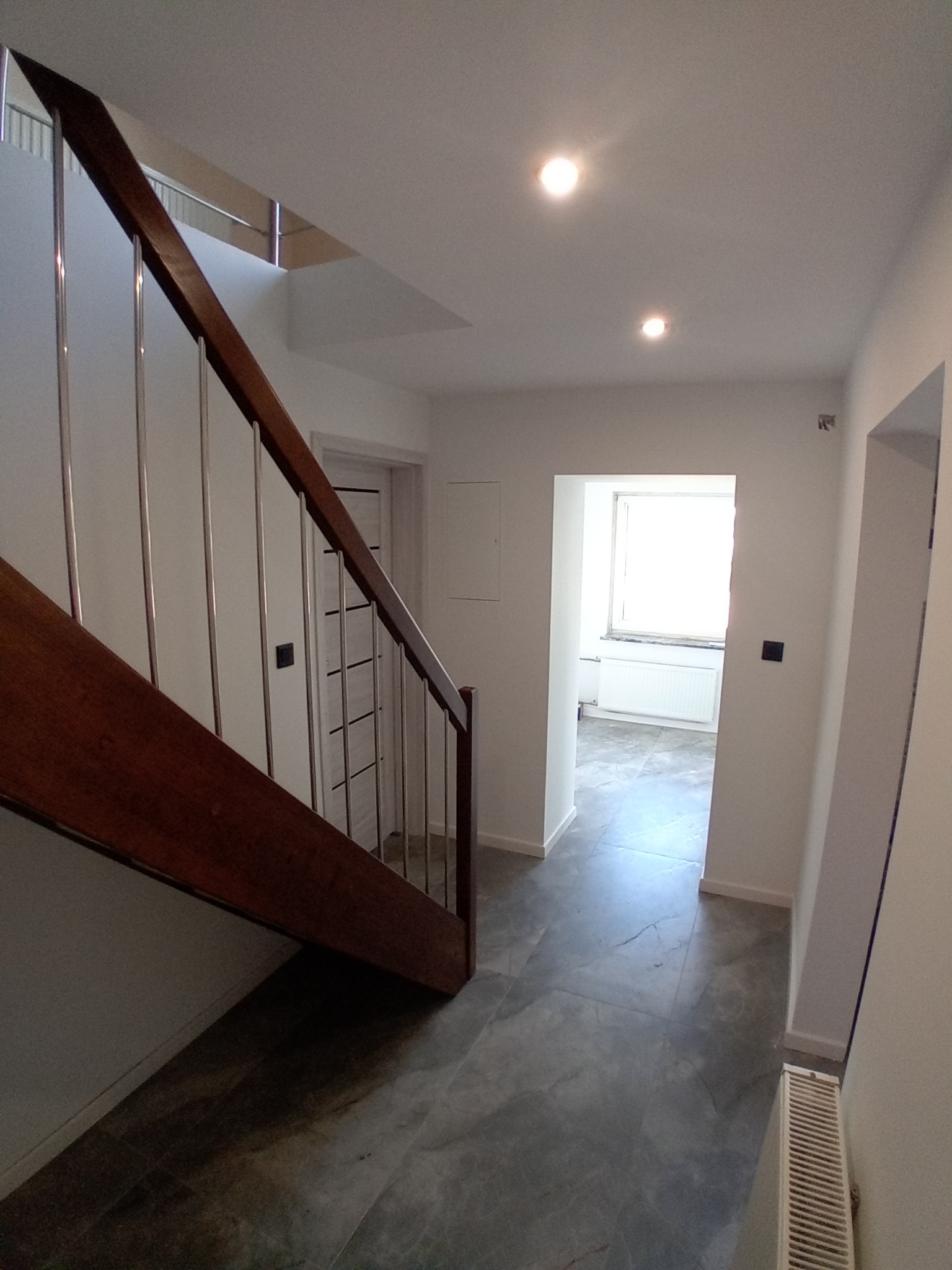 Nowoczesne wnętrze: schody z drewnianą balustradą i metalowymi słupkami, szara podłoga z płytek, białe ściany i drzwi. Widok na jasne pomieszczenie z oknem.