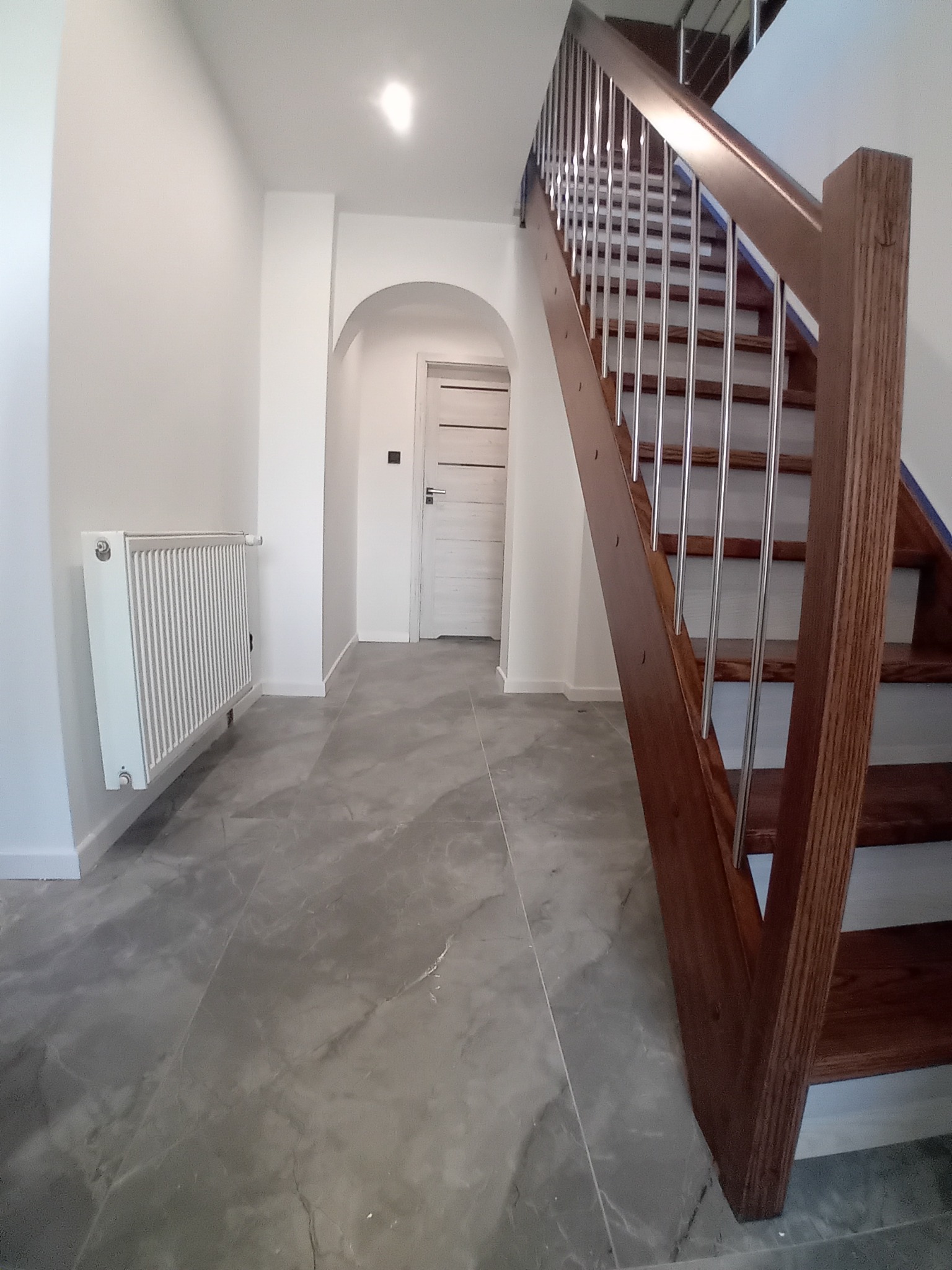 Jasne wnętrze po remoncie: szare płytki podłogowe, białe ściany, grzejnik, drewniane schody z metalowymi barierkami i łukowate przejście z drzwiami.