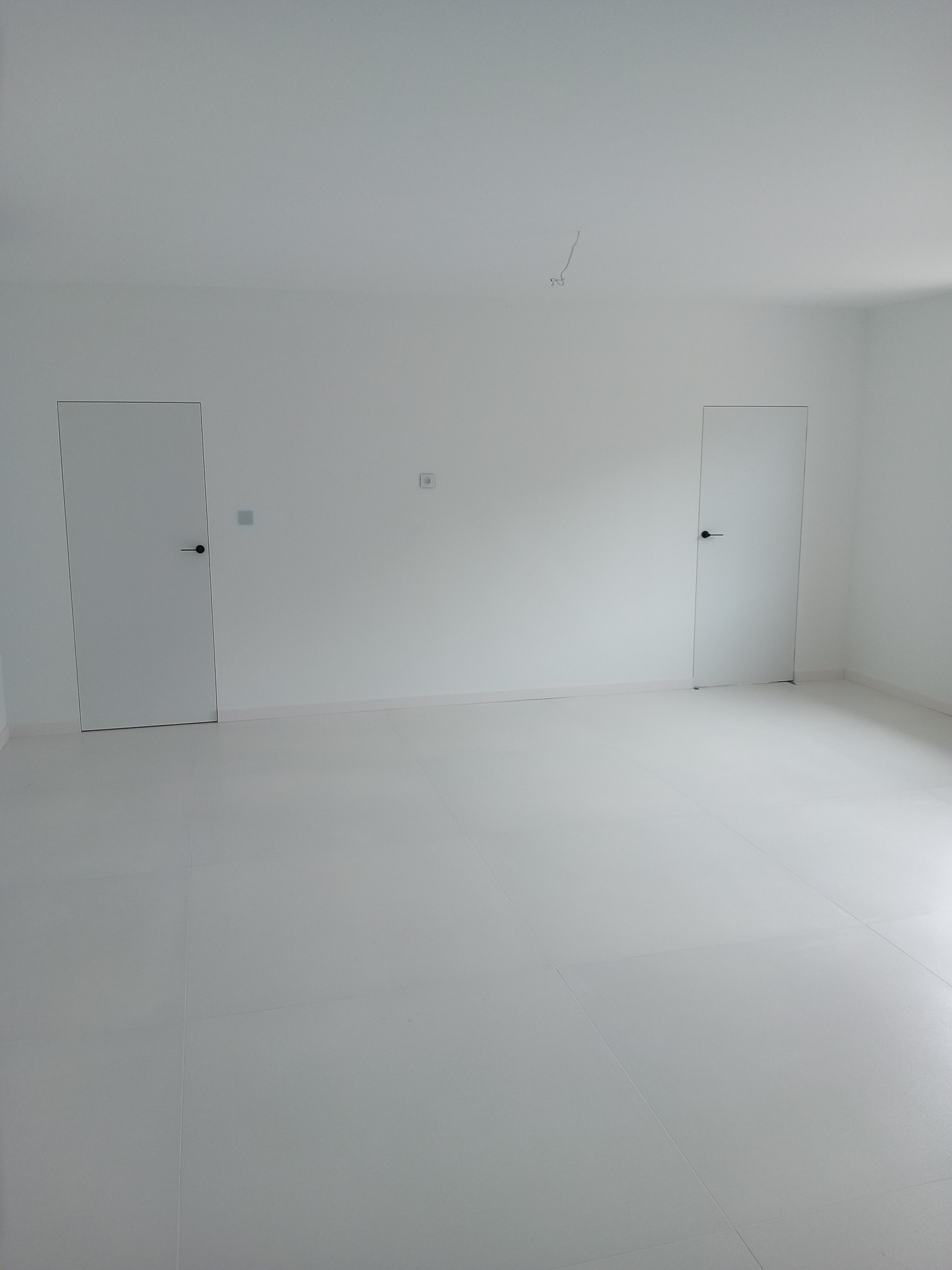 Minimalistyczne, białe wnętrze po remoncie: gładkie ściany, jasna podłoga z płytek, dwie ukryte drzwi z czarnymi klamkami. Nowoczesne wykończenie, surowy styl.