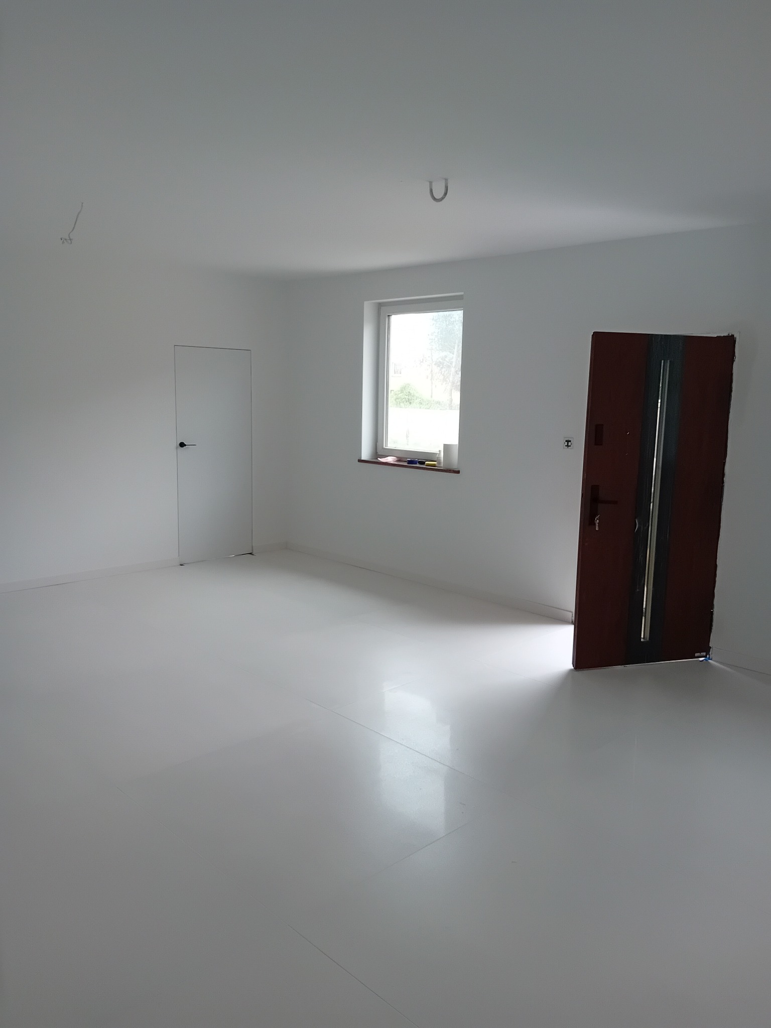 Jasne, puste wnętrze po remoncie: białe ściany, podłoga z połyskiem, okno z parapetem i dwie różne drzwi. Widoczne niedociągnięcia na suficie.