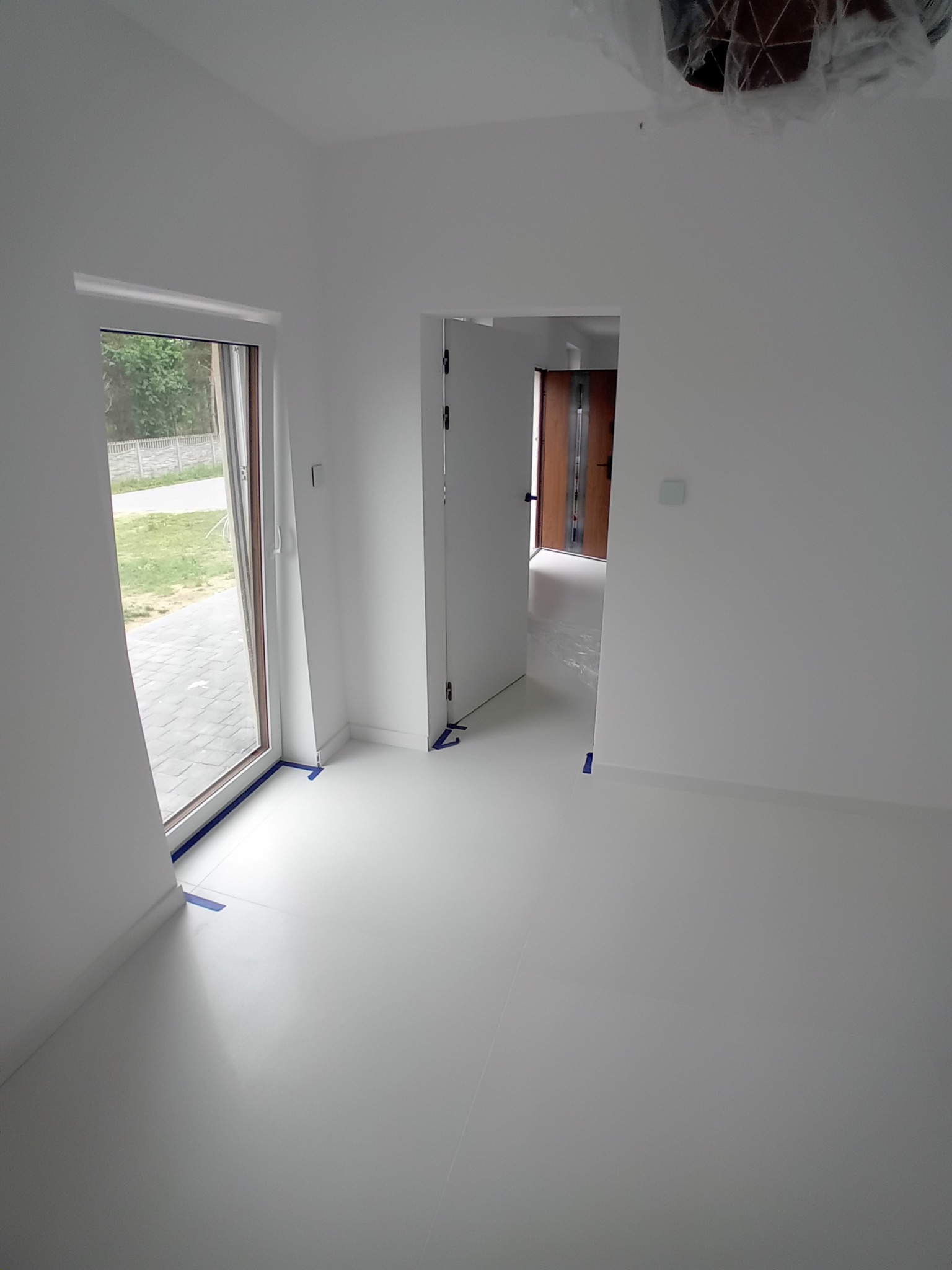 Białe, świeżo pomalowane wnętrze z widokiem na zewnątrz przez okno. Podłoga i ściany jednolicie białe, zabezpieczone taśmą malarską. Widoczny fragment lampy pod sufitem.