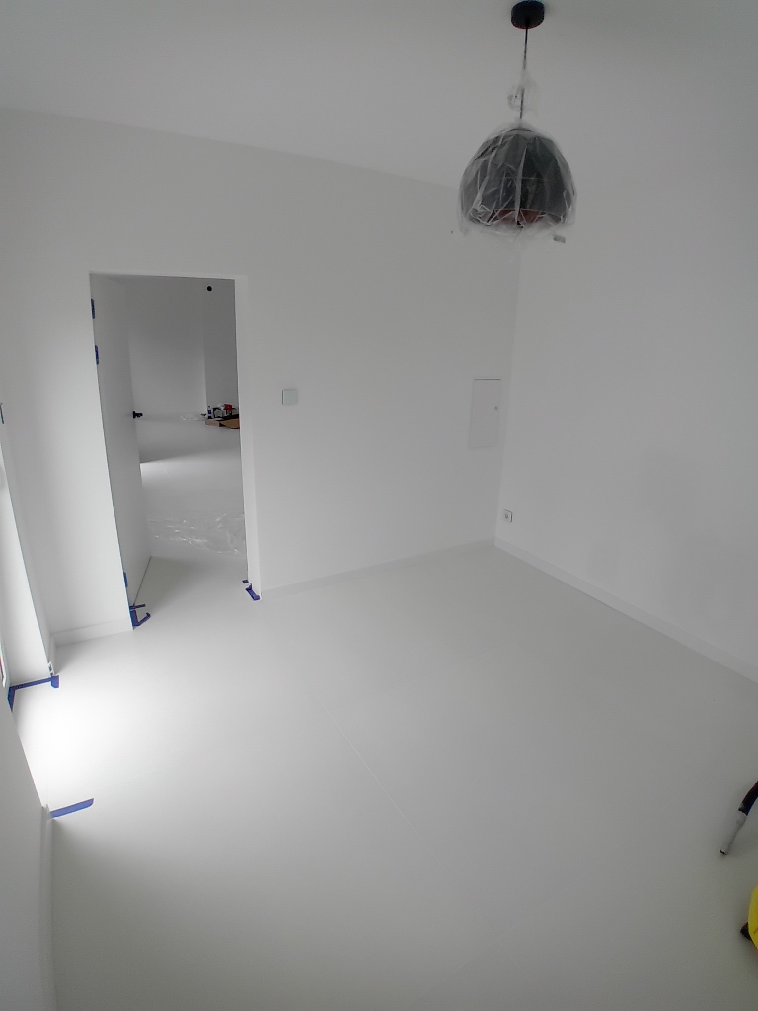 Białe, odświeżone wnętrze pokoju po malowaniu, z zabezpieczoną folią lampą i taśmą malarską przy podłodze, widok na sąsiednie pomieszczenie.