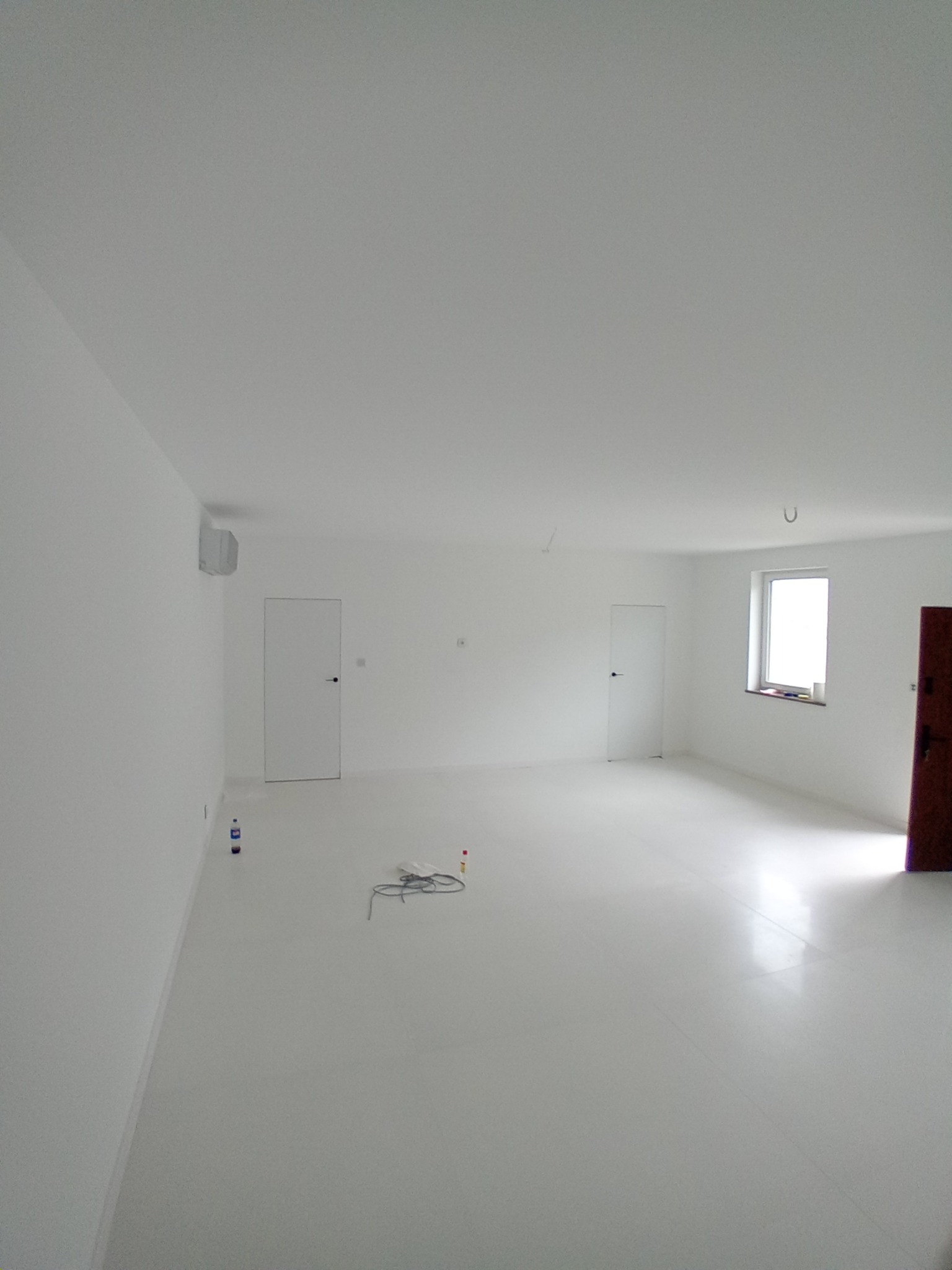 Białe, puste wnętrze po remoncie. Świeżo pomalowane ściany, gładka podłoga, dwie białe drzwi i okno z widokiem na zewnątrz. Minimalistyczny styl, brak mebli, przygotowane do aranżacji.