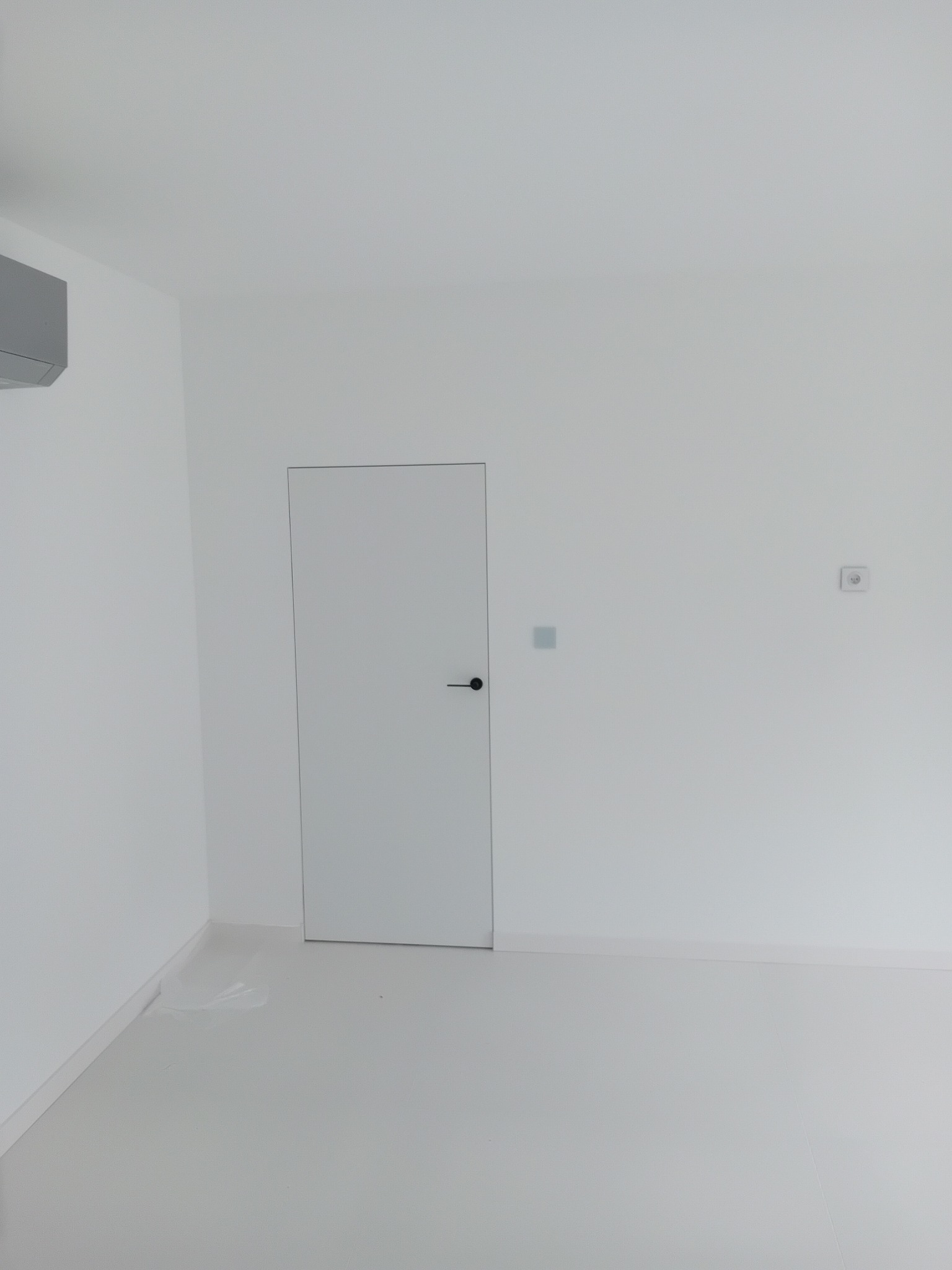 Minimalistyczne wnętrze: białe ściany, podłoga i drzwi z czarną klamką. Widoczny klimatyzator i gniazdko elektryczne. Całość utrzymana w jasnej tonacji.