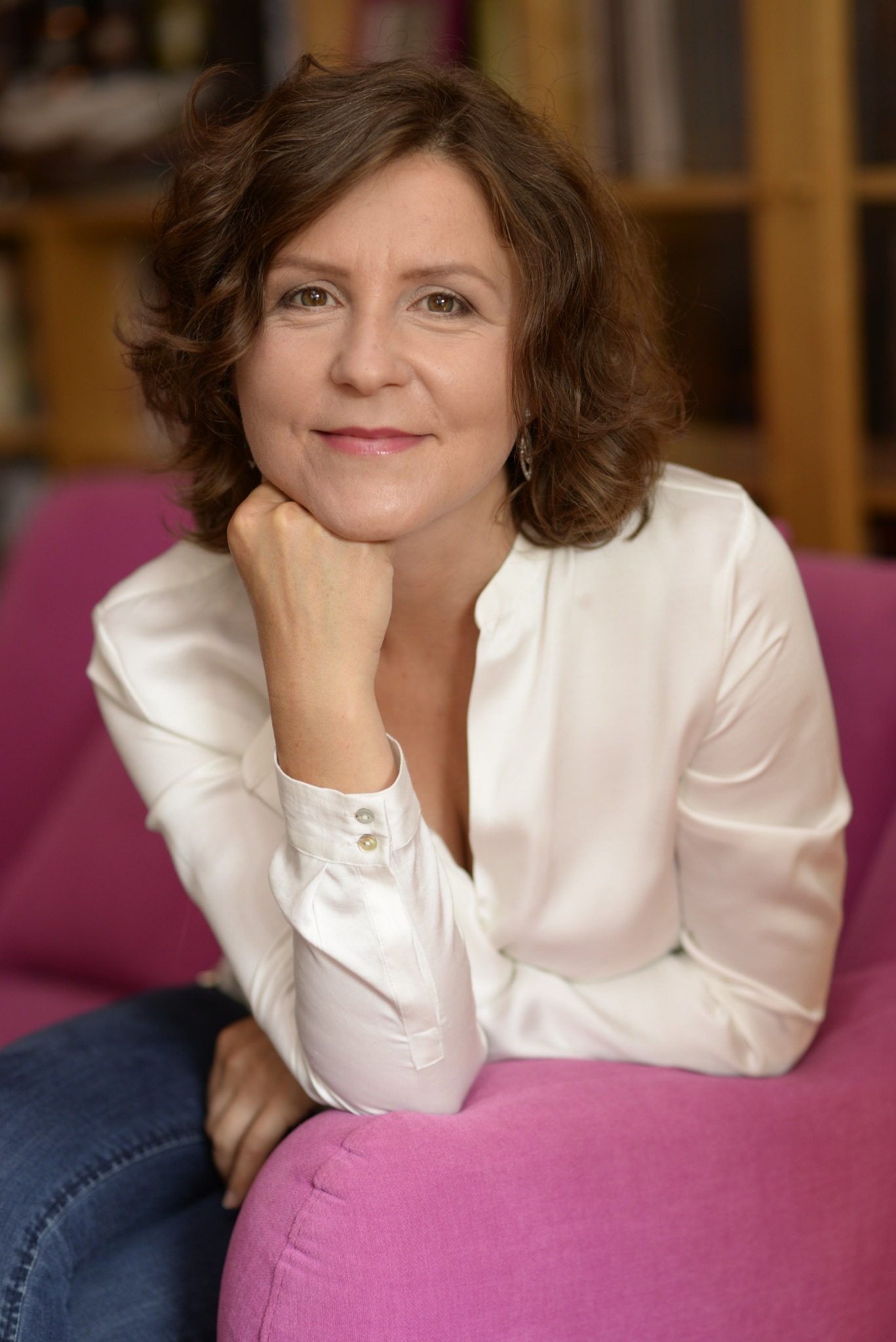 Portret kobiety w białej bluzce siedzącej na różowej sofie, z biblioteczką w tle, spoglądającej w obiektyw z delikatnym uśmiechem.