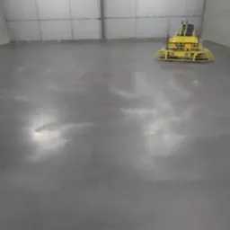 Świeżo wylana, szara posadzka w hali przemysłowej, z żółtą maszyną do zacierania betonu w tle.