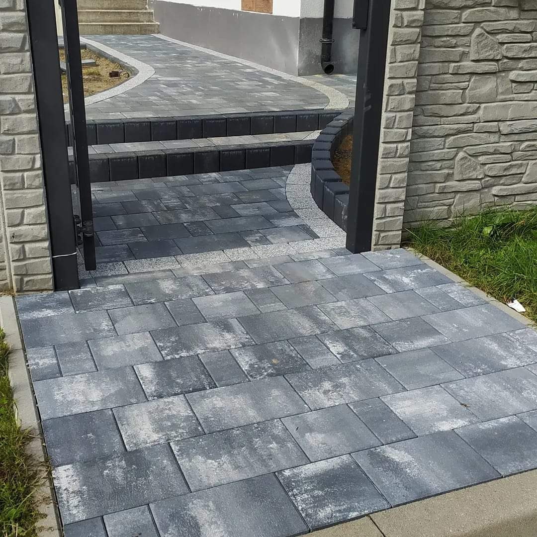 Ułożona z szarych kostek ścieżka prowadząca przez otwartą, czarną bramę do domu z kamienną elewacją i widocznymi schodami.