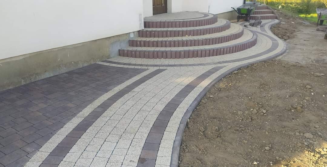 Ułożona ścieżka z kostki brukowej prowadząca do schodów wejściowych domu, z widocznymi elementami w kolorze szarym i brązowym, oraz częściowo przygotowanym terenem do dalszych prac.