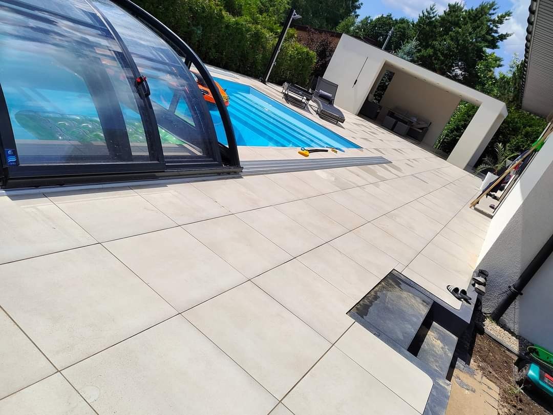 Jasne, duże płyty tarasowe otaczają basen z częściowo otwartym zadaszeniem i przesuwnymi drzwiami; w tle minimalistyczna altana z meblami ogrodowymi.