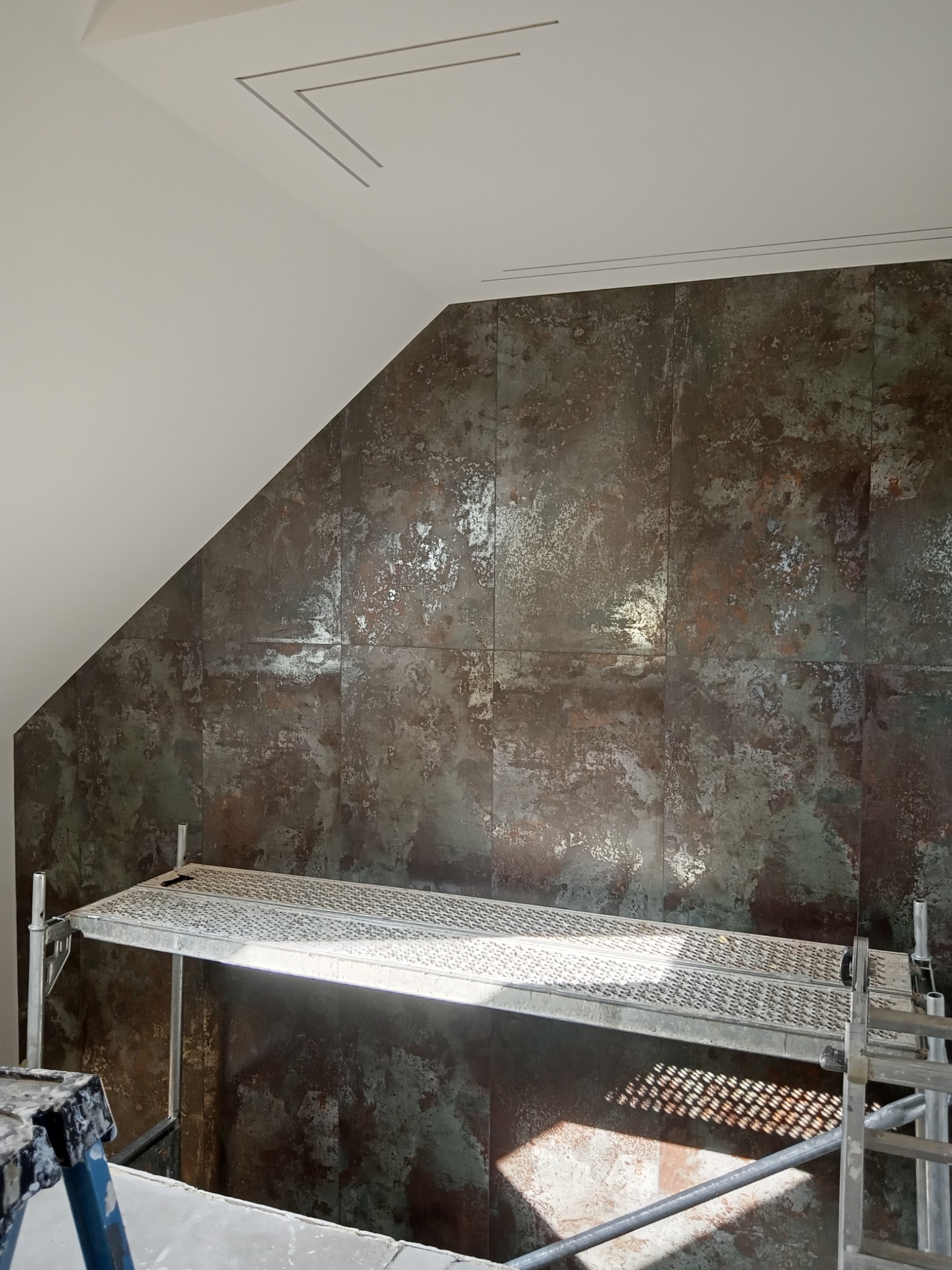 Ściana wyłożona płytami imitującymi rdzę, widoczny fragment rusztowania i drabiny malarskiej w pomieszczeniu ze skośnym sufitem.