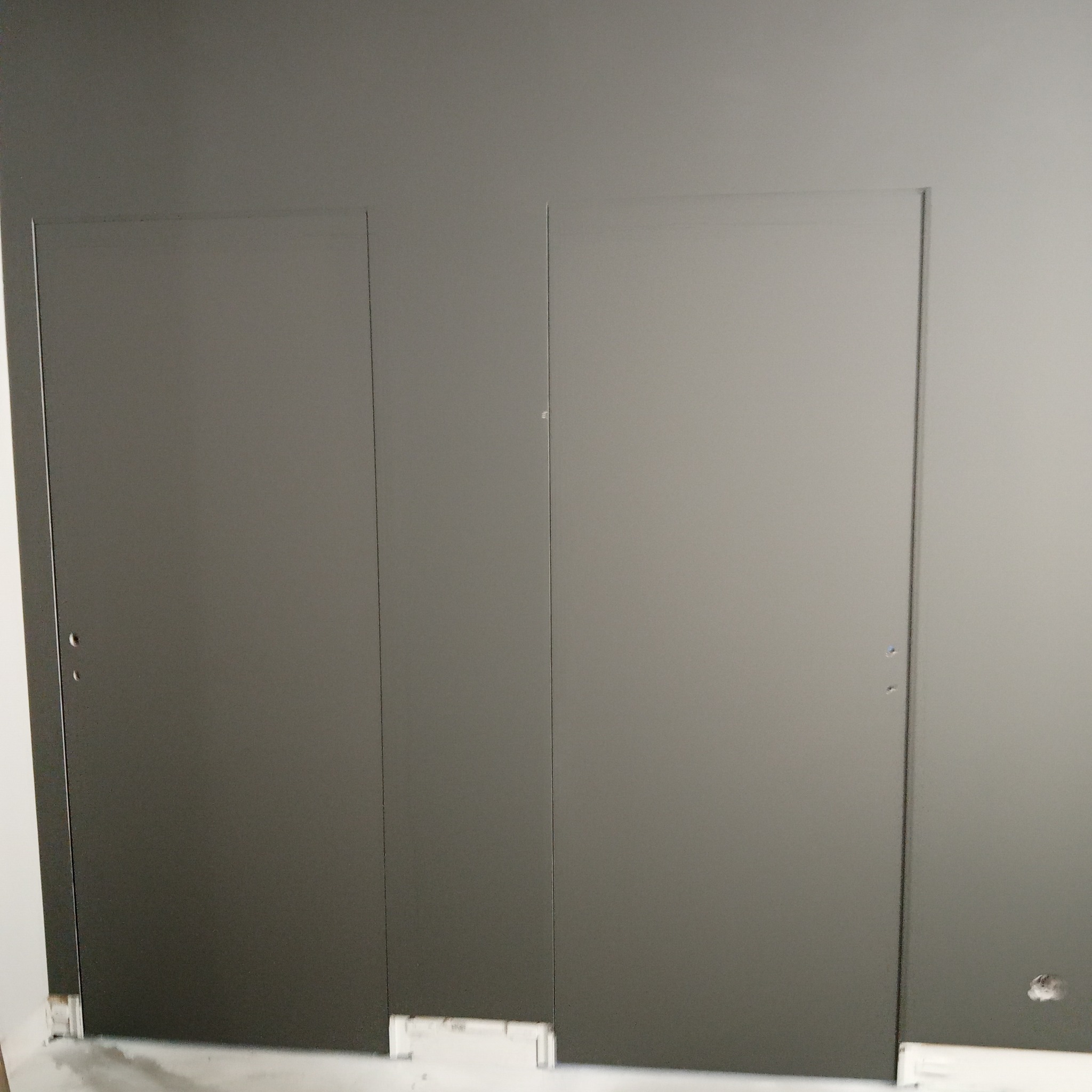 Szara ściana z dwiema ukrytymi drzwiczkami rewizyjnymi, widoczne niedoskonałości wykończenia przy podłodze.