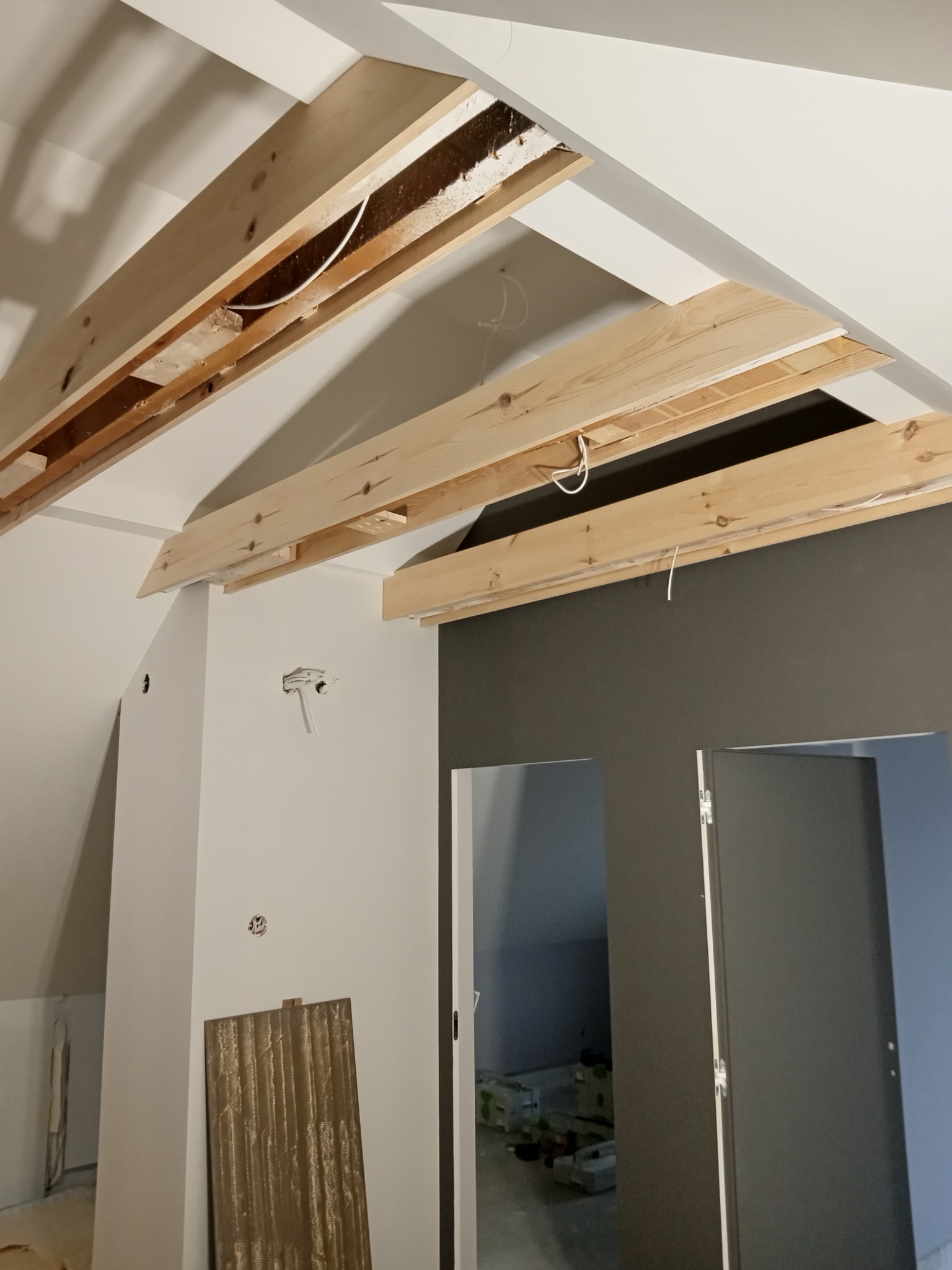 Wnętrze poddasza w trakcie prac wykończeniowych, widoczne drewniane belki konstrukcyjne sufitu podwieszanego, instalacja elektryczna, fragment ściany pomalowanej na szaro i drzwi.