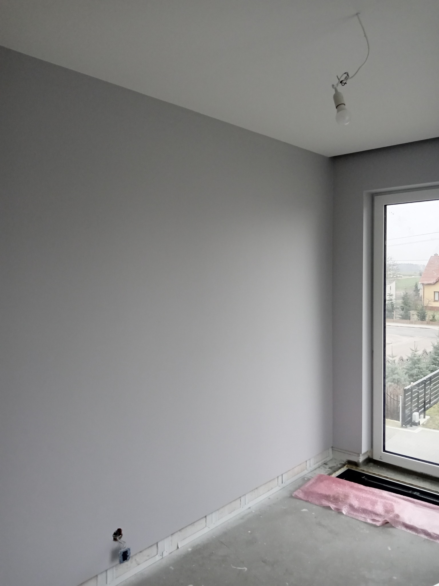 Wnętrze pokoju w szarych odcieniach z gładkimi ścianami, widoczne przygotowania do wykończenia podłogi, okno z widokiem na dom i ogród, oraz prowizoryczne oświetlenie.