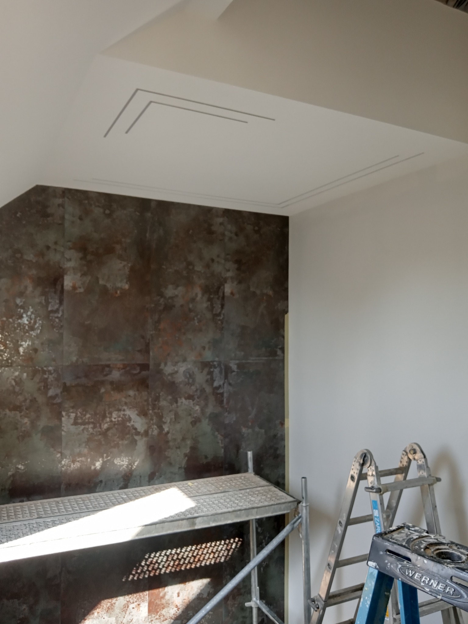 Wnętrze w trakcie prac wykończeniowych: ściana z płytek imitujących rdzę, widoczny fragment rusztowania i drabina Werner, sufit z dekoracyjną listwą.