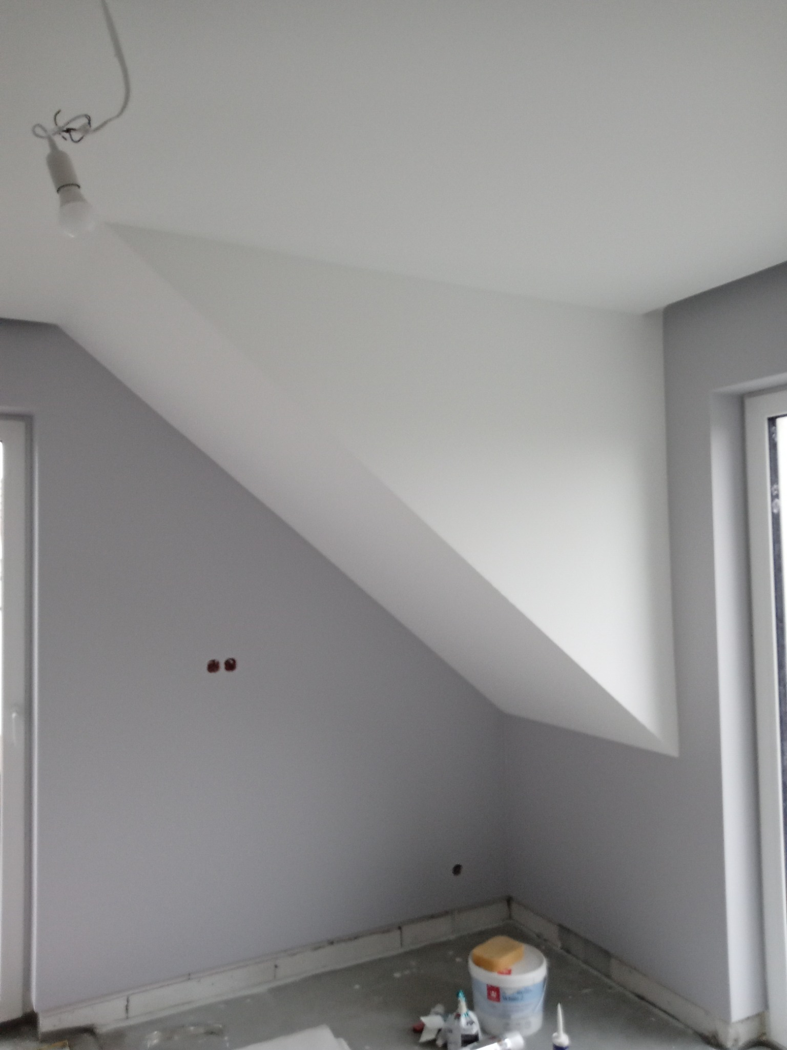 Pokój na poddaszu w trakcie malowania: szare ściany, biały sufit ze skosem, widoczne puszki z farbą i narzędzia malarskie na podłodze, surowa żarówka na kablu.